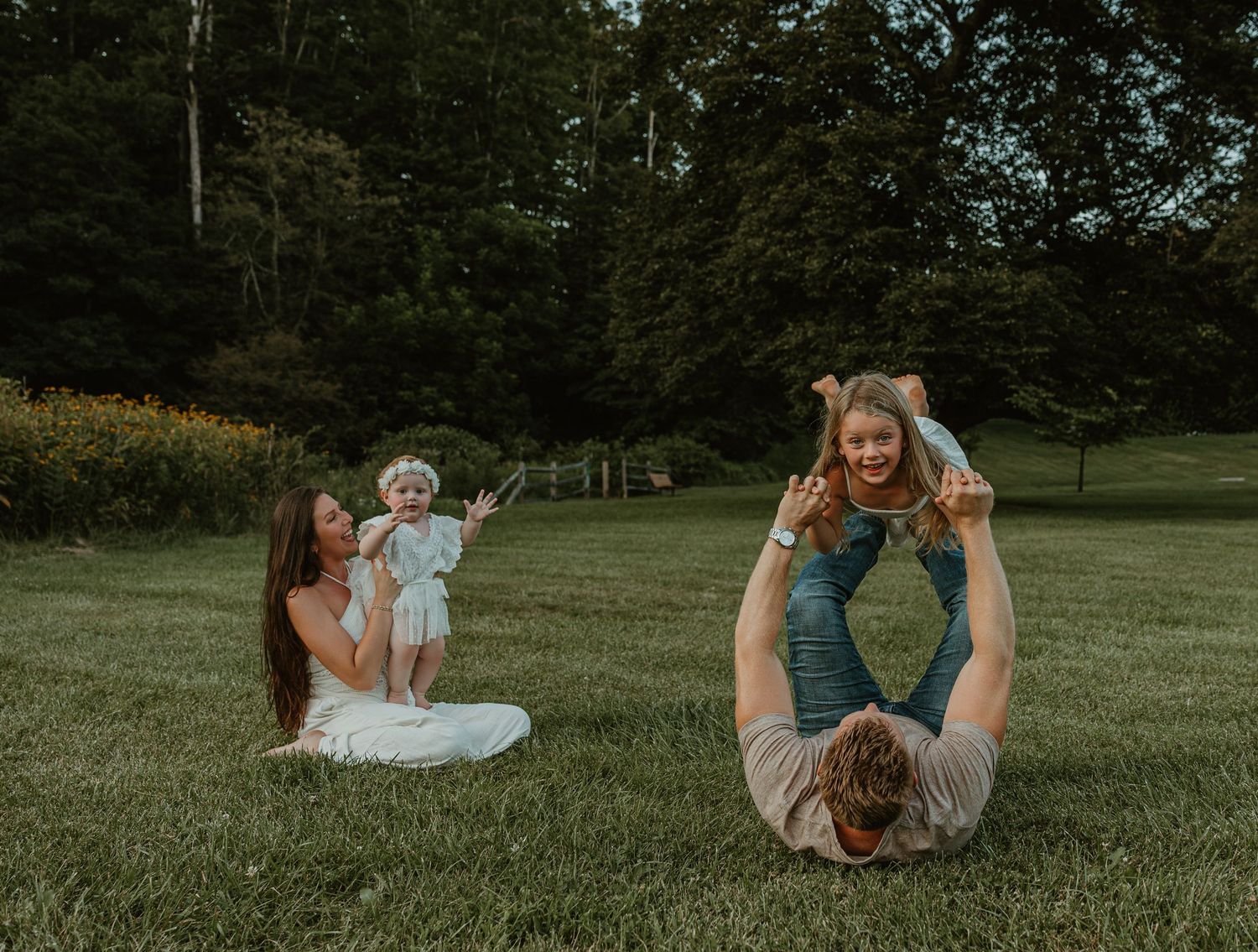 Family playing outdoors on green grass, mother with baby and father lifting toddler in air, surrounded by trees.