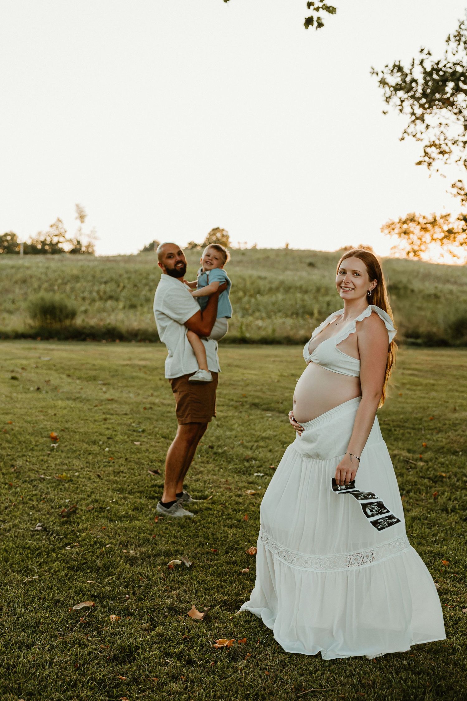 Pregnant woman in white dress plays with toddler in grassy field at sunset.