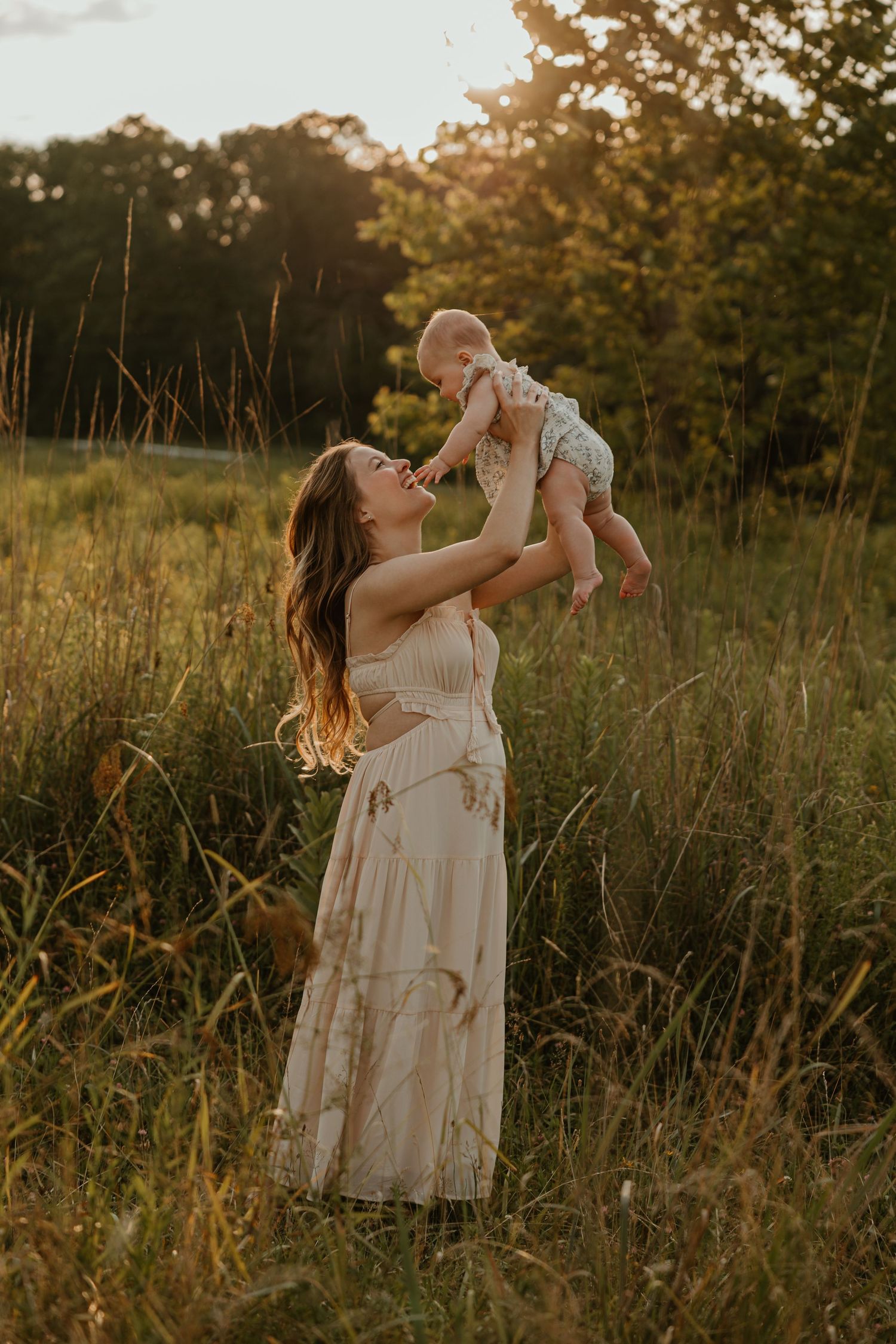 A mother in a flowy dress lifts her baby in a sunlit meadow at golden hour.