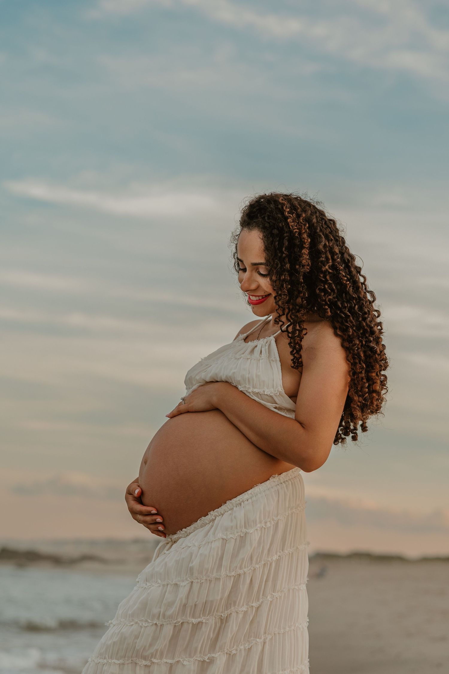 Pregnant woman with curly hair in white dress cradling her baby bump on a beach at sunset.