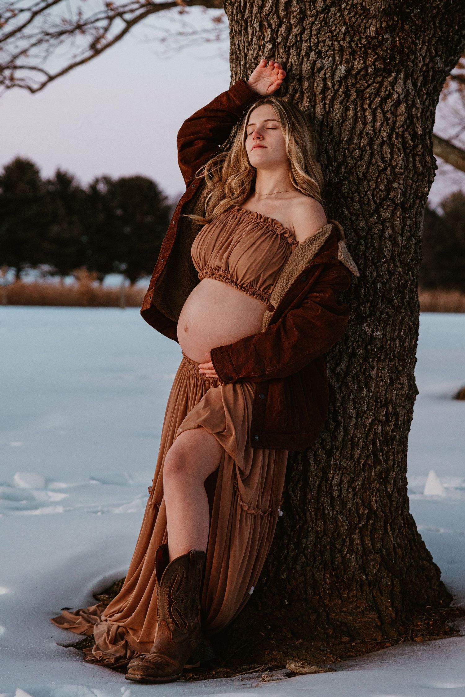Dramatic maternity portrait against a tree trunk with snow and warm sunset lighting.