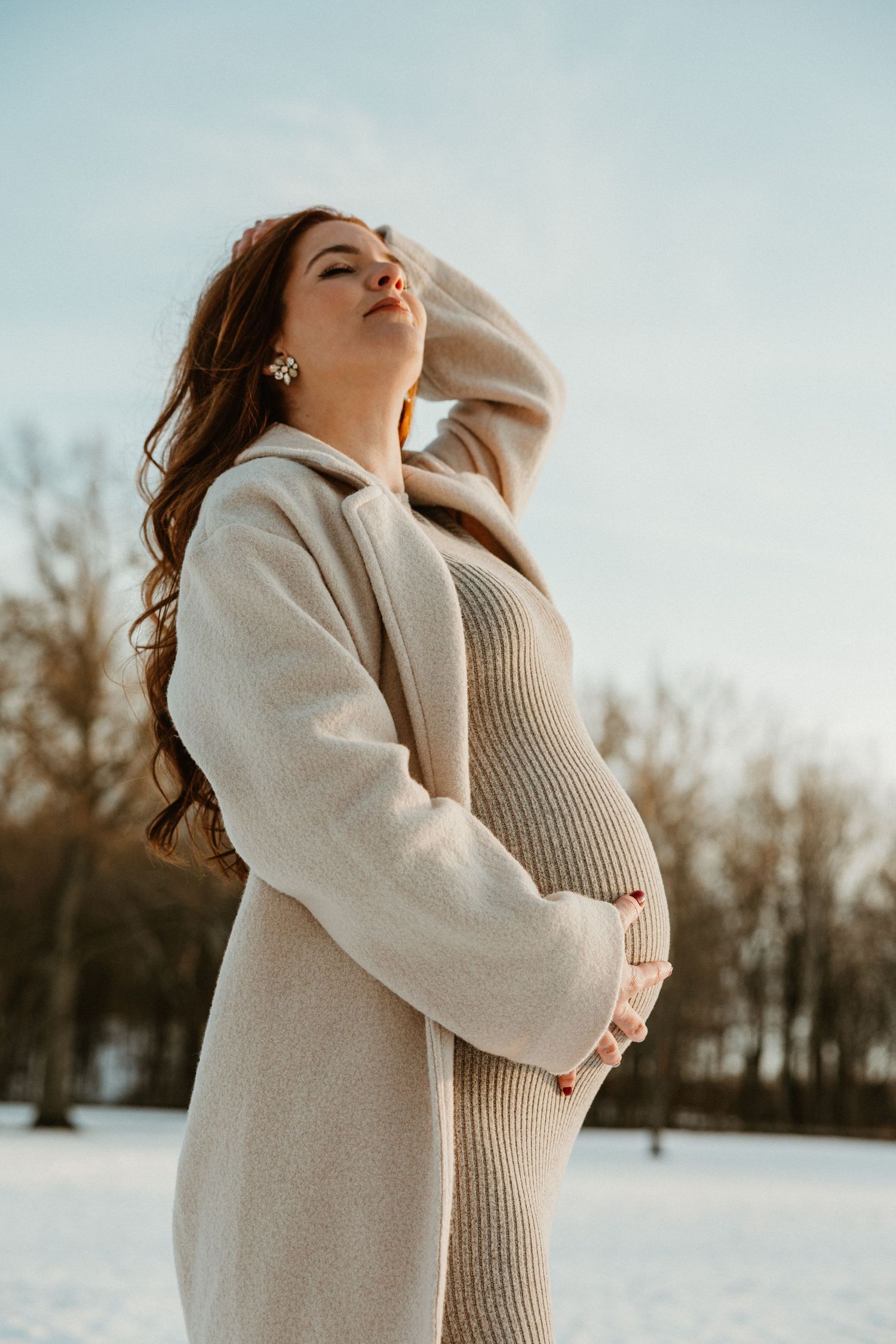 Winter maternity portrait featuring woman in beige coat against snow-covered scenery.
