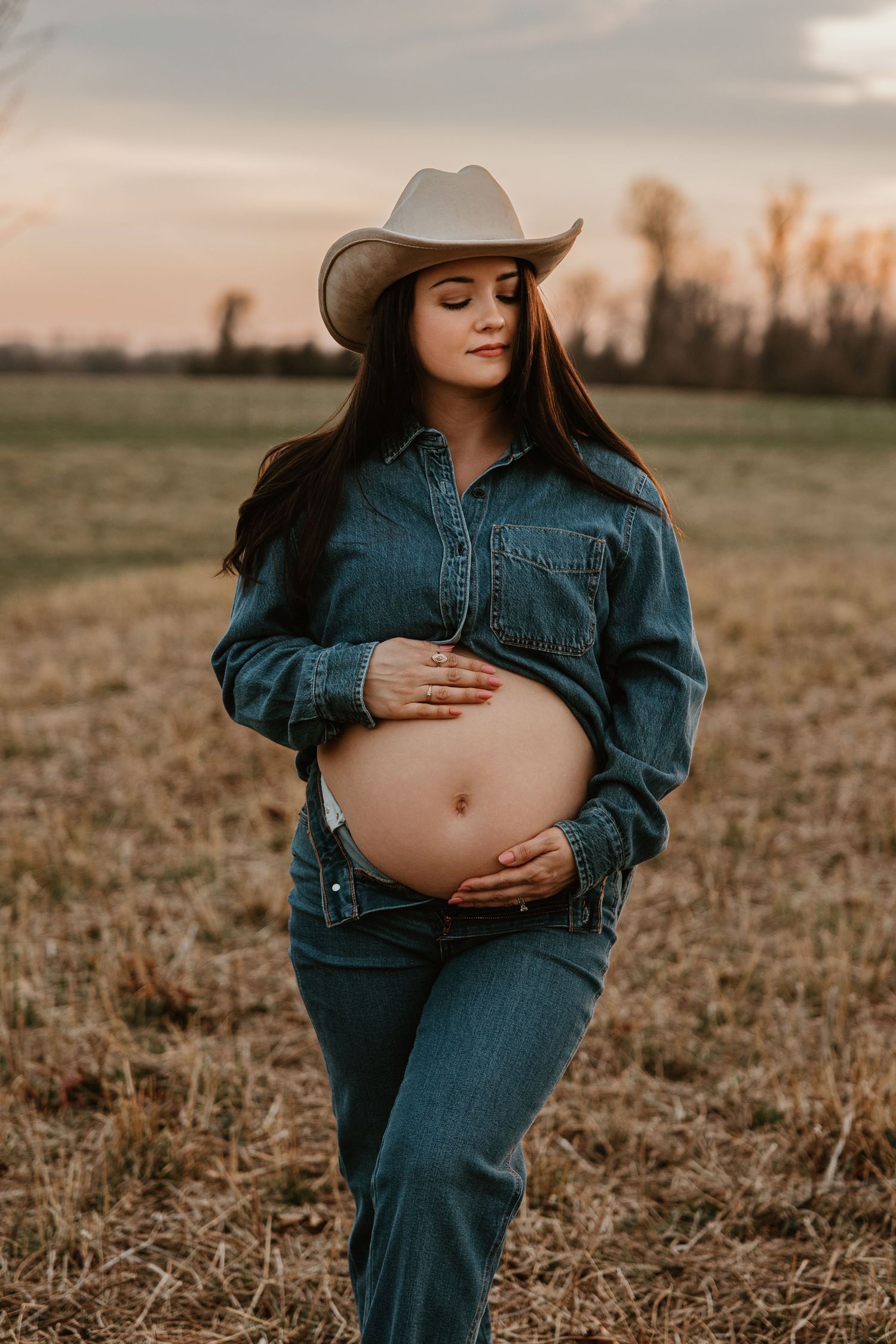 Pregnant woman in denim outfit and cowboy hat cradling baby bump in a golden field at sunset.