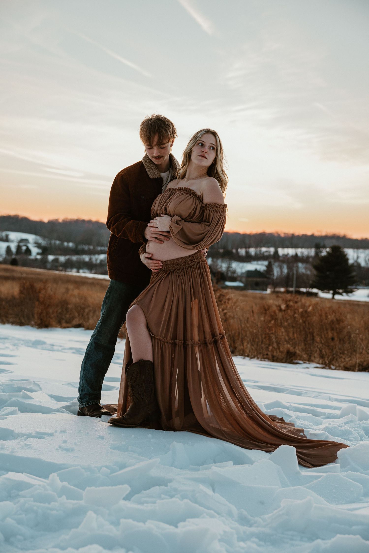 Romantic maternity photo of couple in brown dress standing in snowy field at sunset.