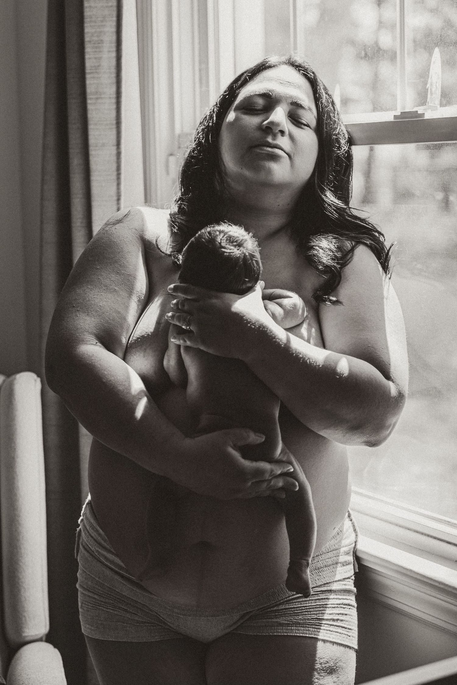 Black and white portrait of mother holding newborn baby near window in emotional postpartum moment.