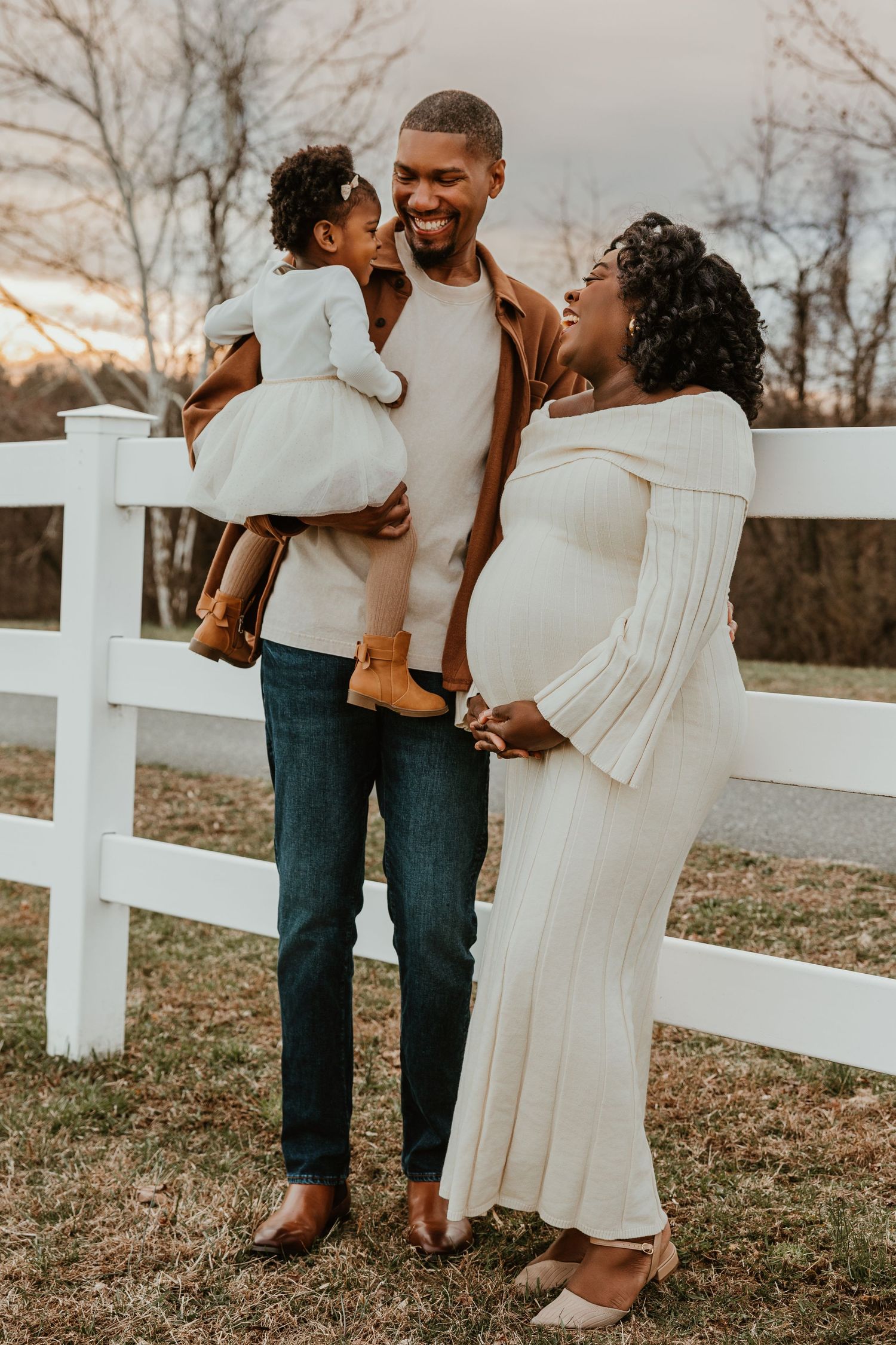 Happy Black family maternity photo with pregnant mother in cream dress, father holding toddler by white fence.