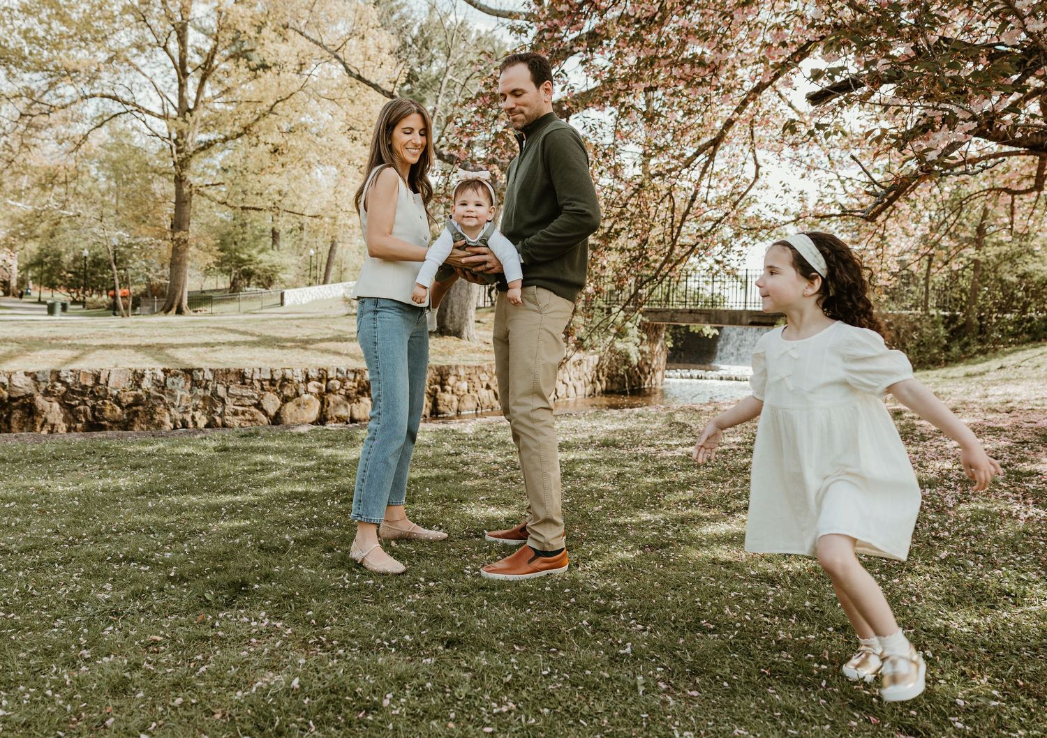 Family of four enjoying a sunny day outdoors, parents holding toddler while young girl twirls in white dress.