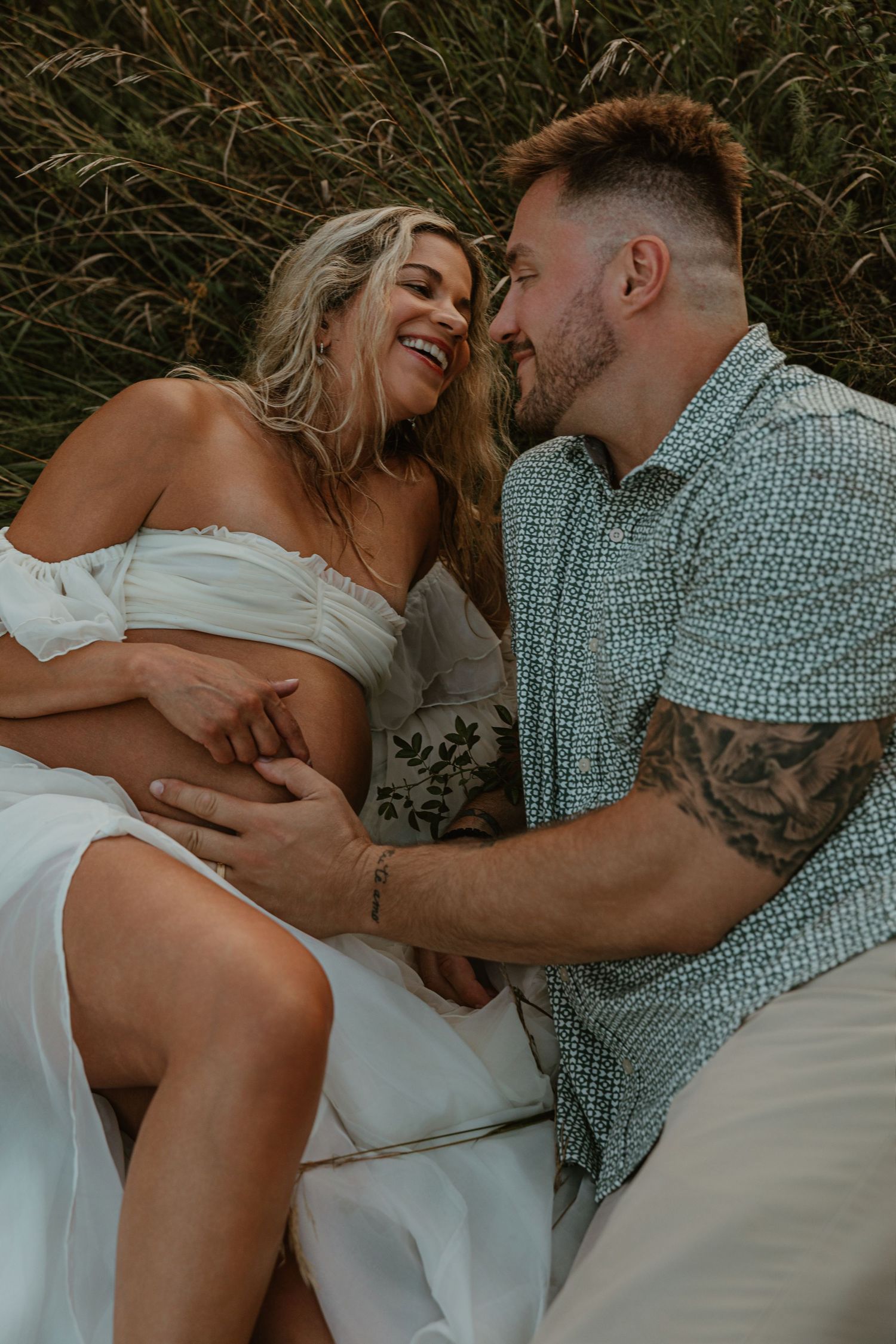 Pregnant woman in white dress laughs with tattooed partner outdoors in tall grass during maternity photo session.