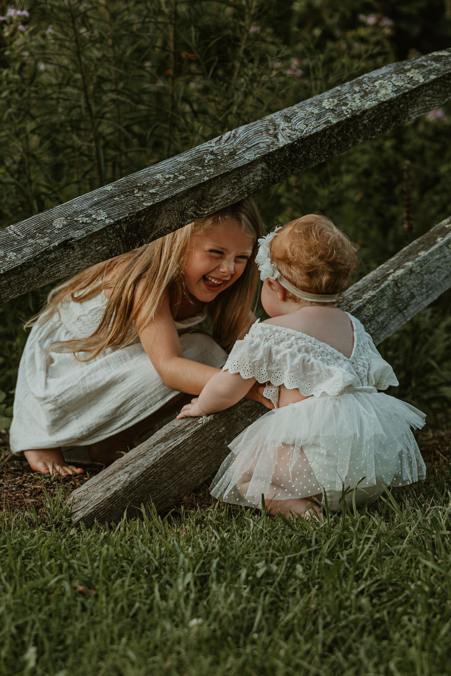 Two young girls in white dresses laugh together near a rustic wooden fence in a lush green garden.