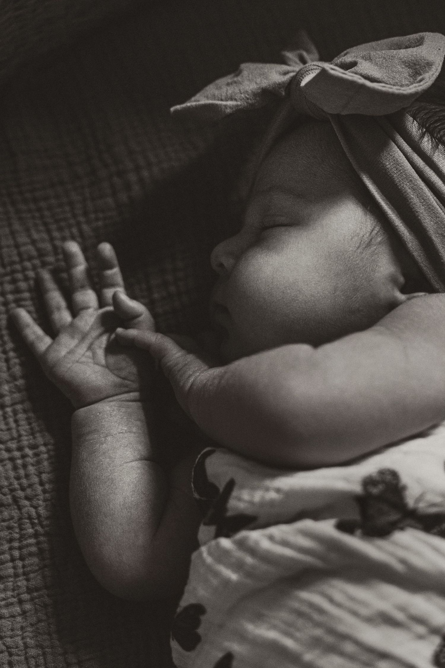 Black and white photo of a sleeping newborn baby wearing a bow headband, with tiny hands visible.