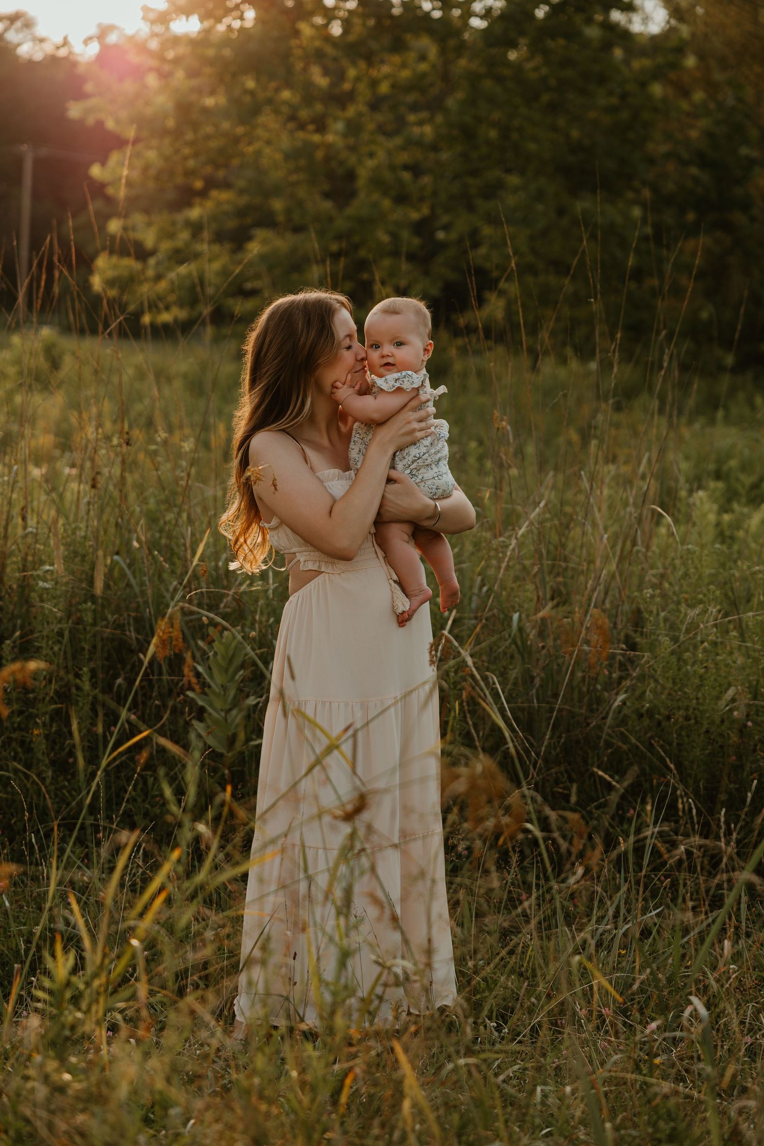 A mother in a white maxi dress holds her baby in a sunlit meadow at golden hour.