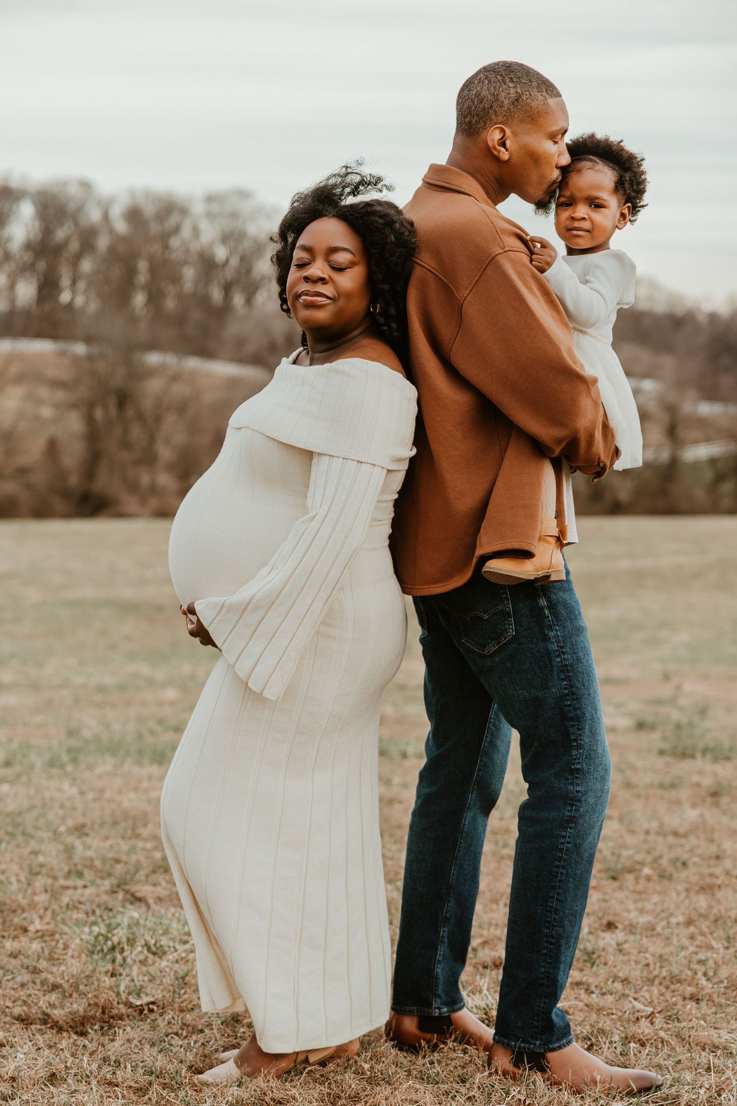 A pregnant woman in a white dress stands with her partner holding their toddler during an outdoor maternity photo session.