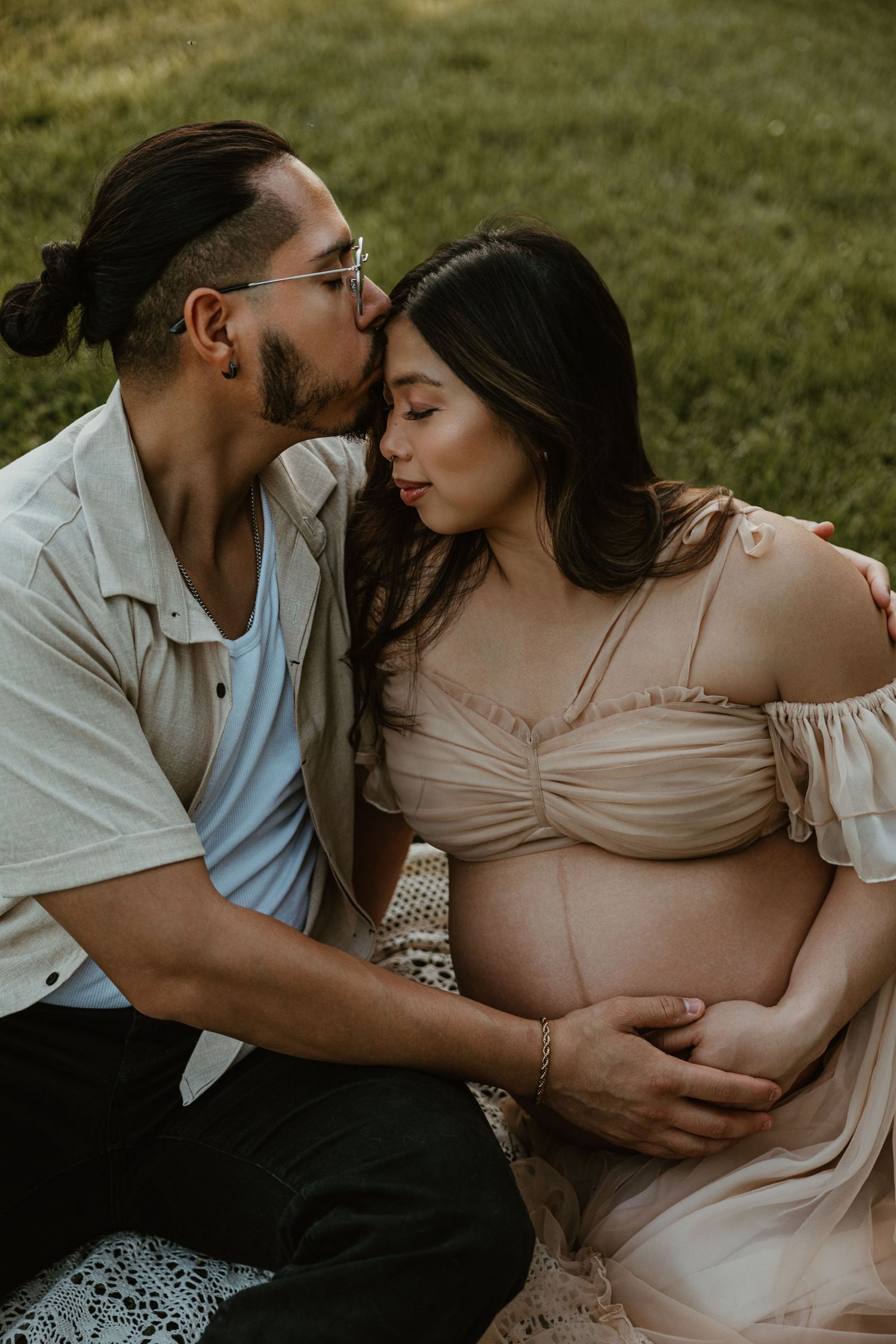 A man kisses a pregnant woman on the cheek as she cradles her baby bump while sitting outdoors on grass.