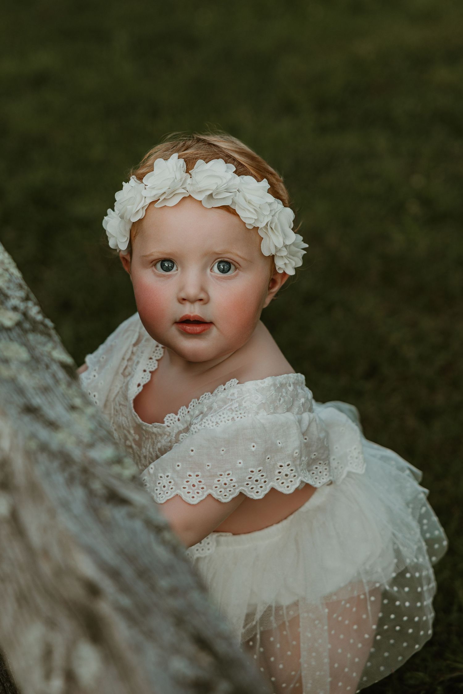 Toddler girl in white lace dress and floral headband poses outdoors near a tree trunk on green grass.