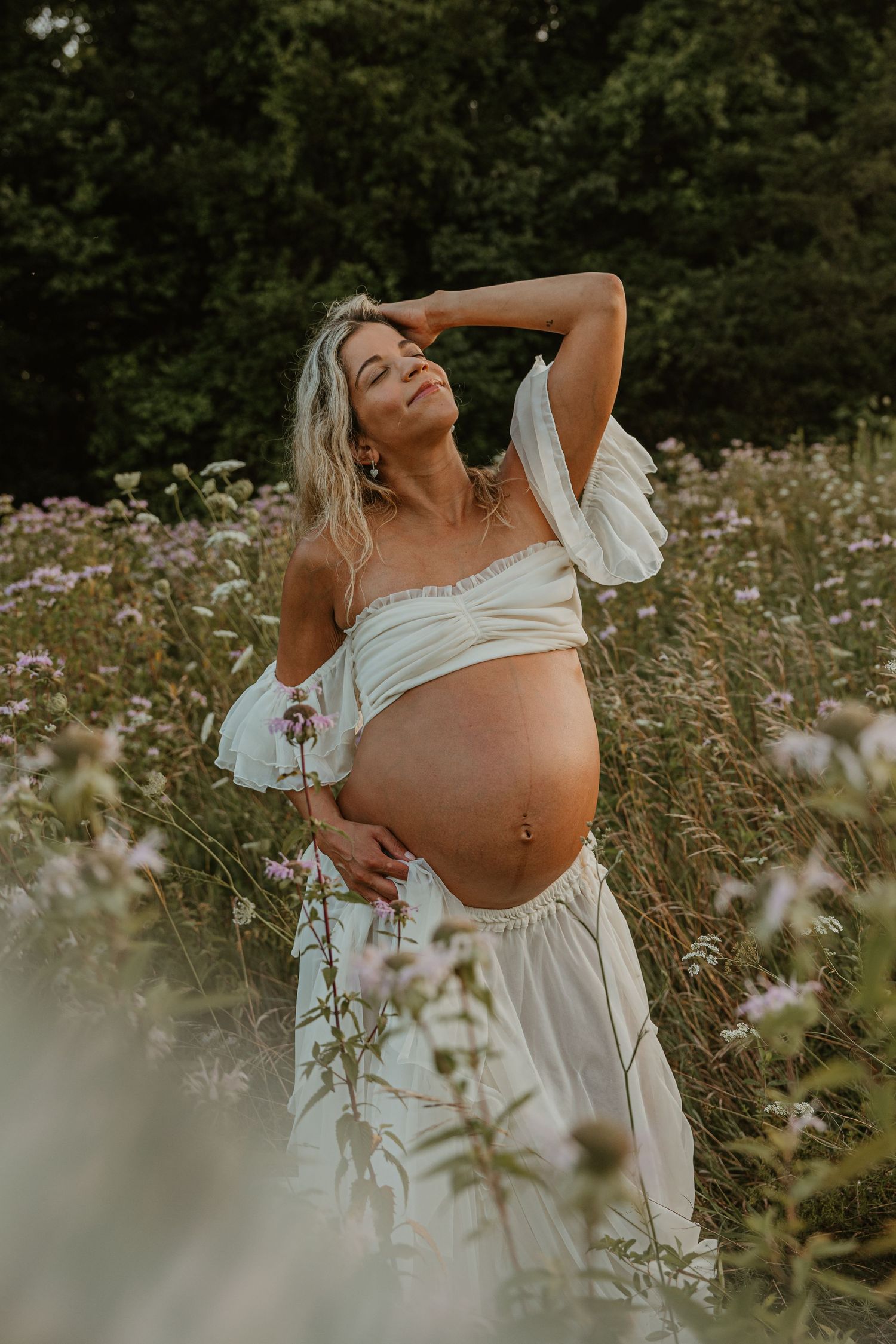 Pregnant woman in white crop top and skirt posing in a wildflower meadow, cradling her baby bump outdoors.