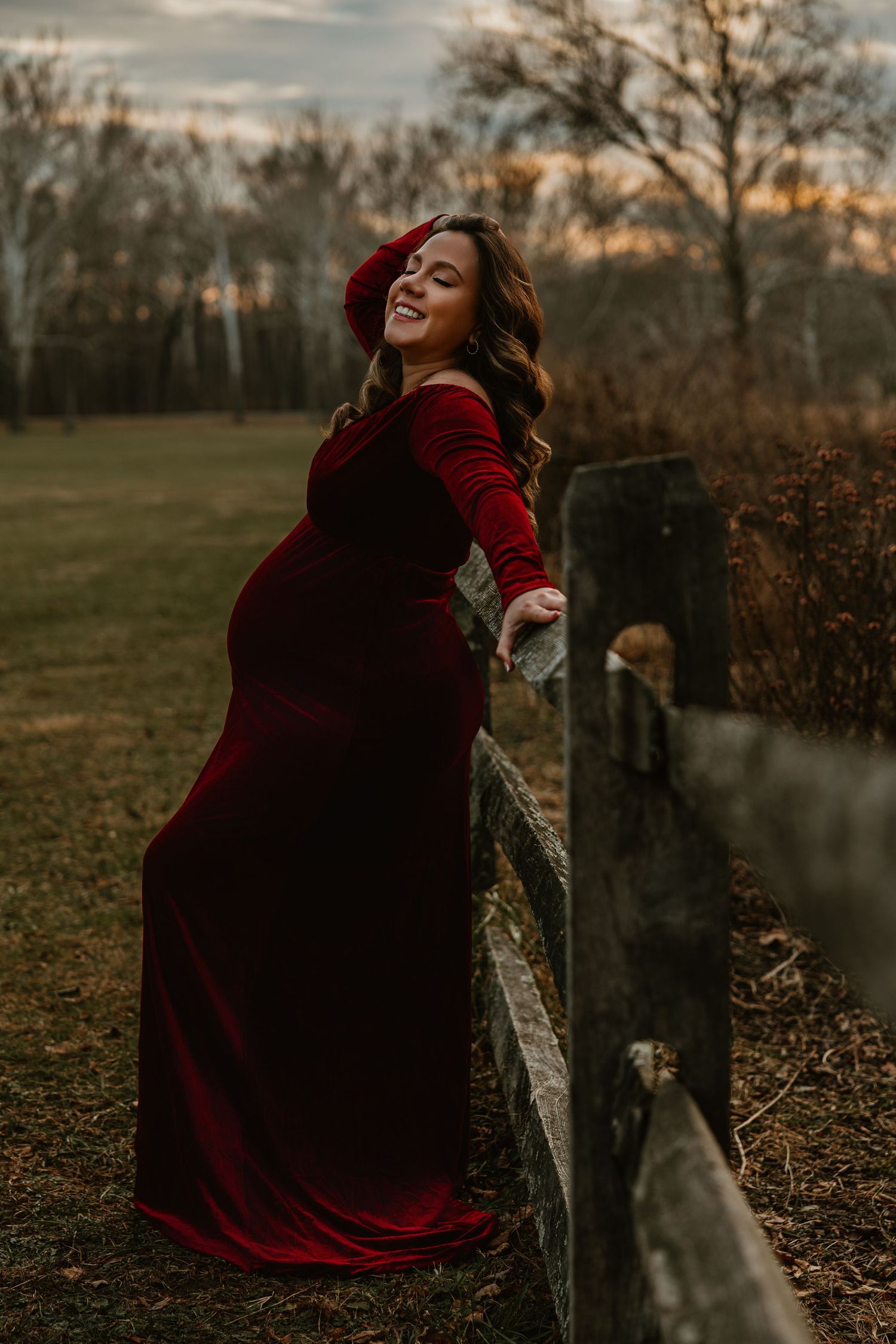 A person in a burgundy velvet dress poses dramatically against an autumn landscape at sunset.