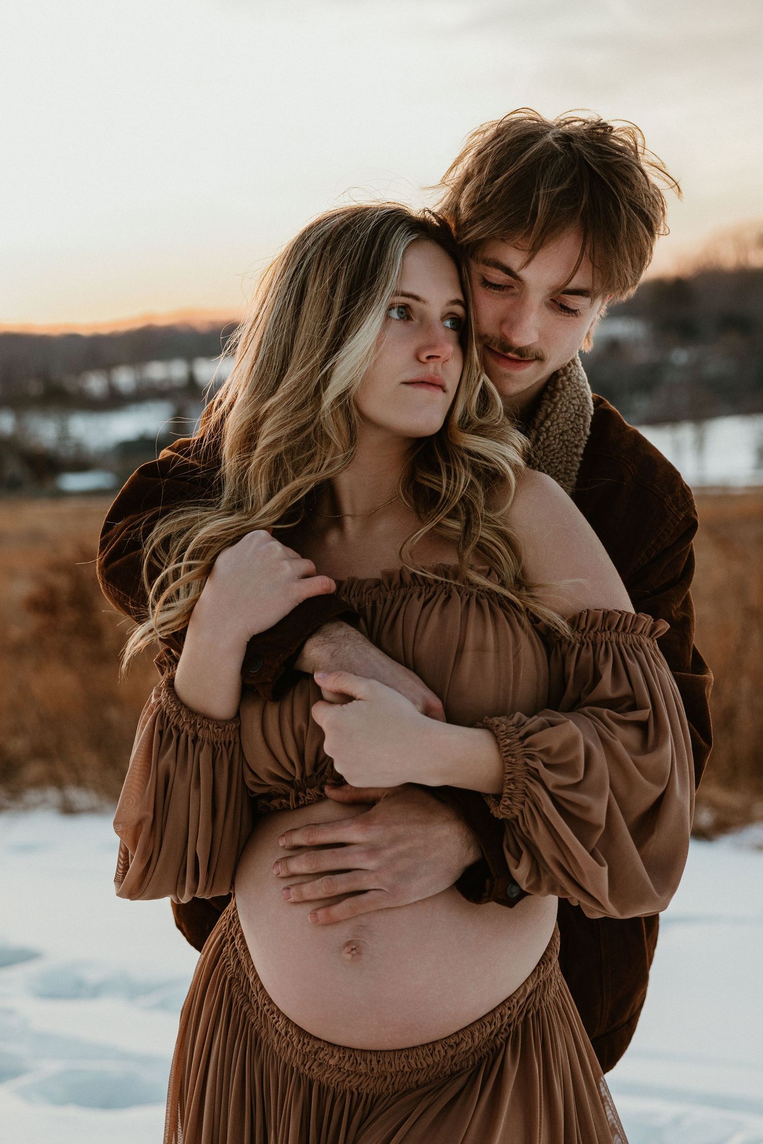 Intimate winter maternity portrait of couple in brown dress against snowy field at golden hour.