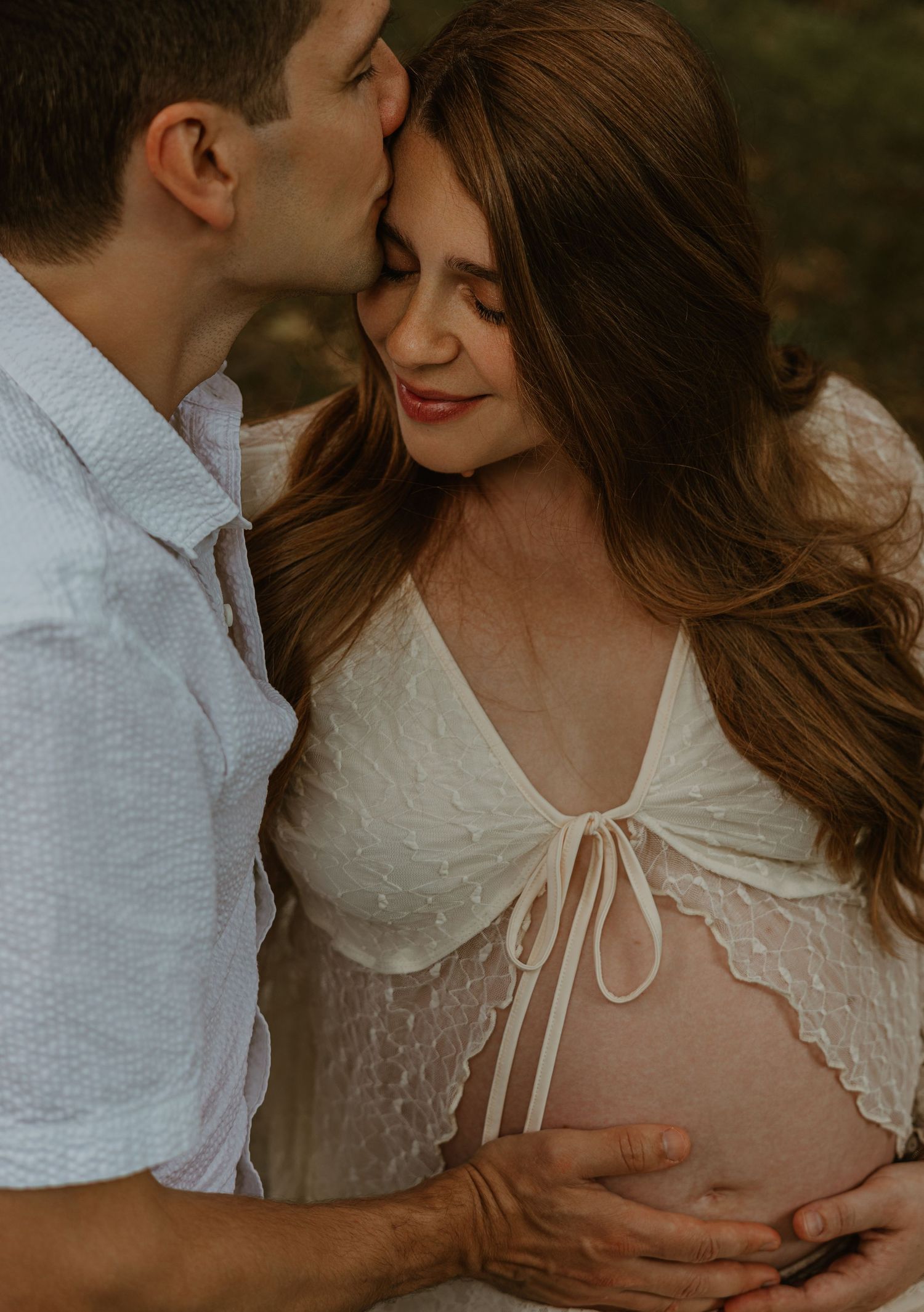 A tender maternity photo of a couple, man kissing woman's head as they cradle her pregnant belly outdoors.