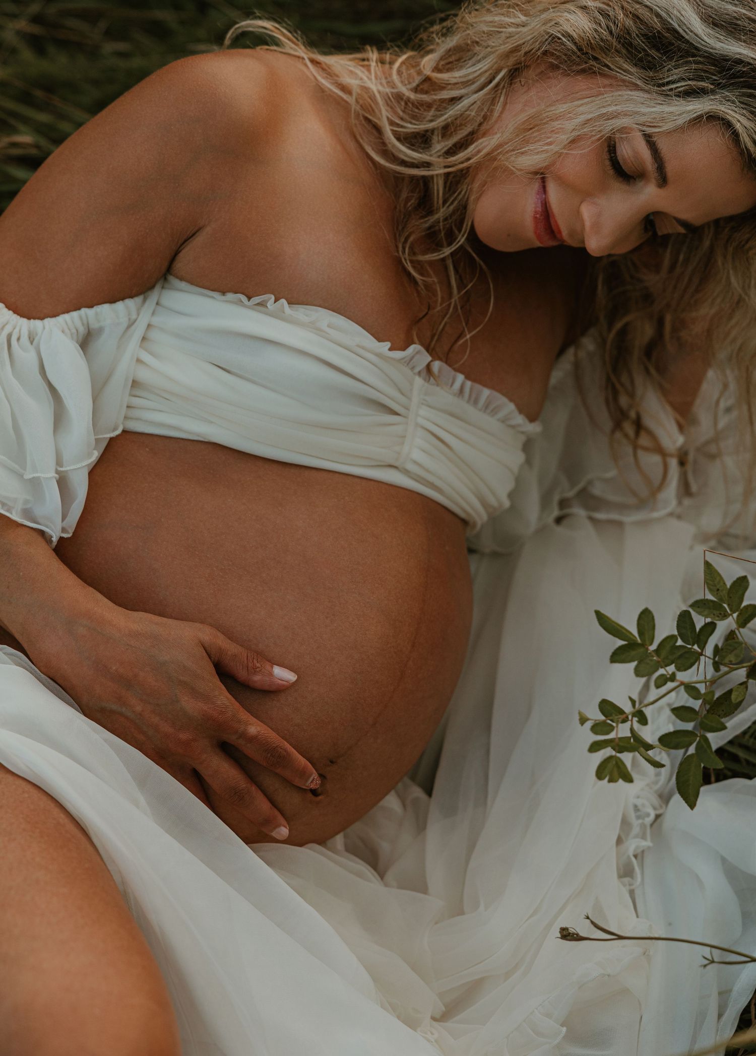 Pregnant woman in white off-shoulder dress cradling baby bump outdoors.