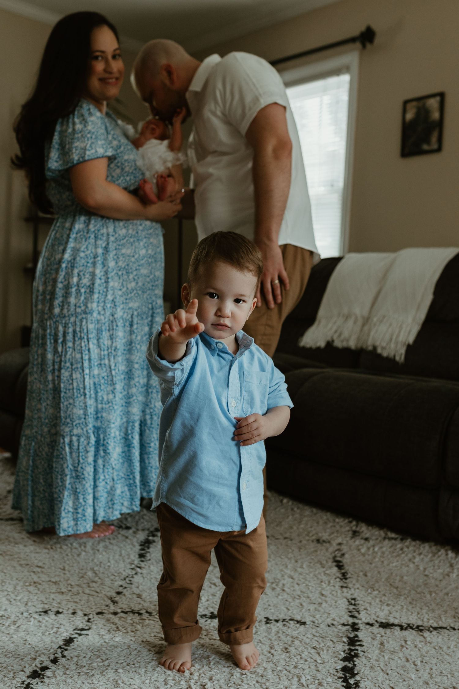 A toddler in blue and brown stands on a white carpet while family members watch from behind.