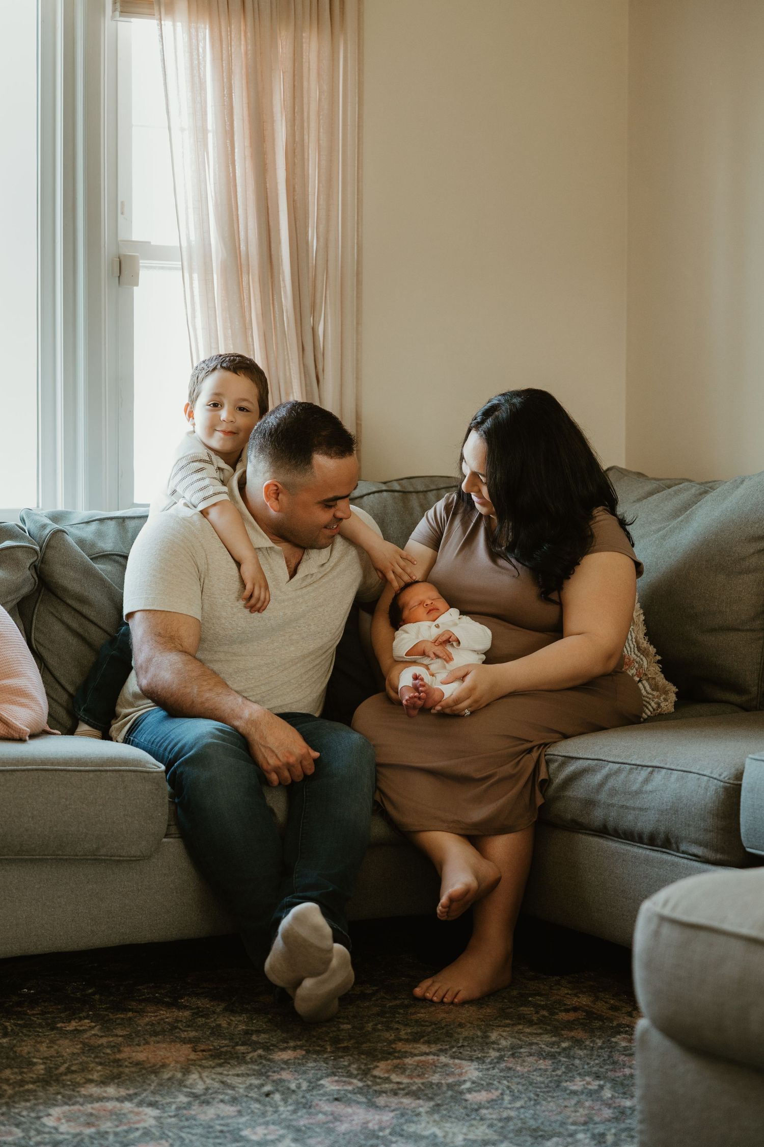 Family of four with newborn baby sharing tender moment together on gray sectional sofa.