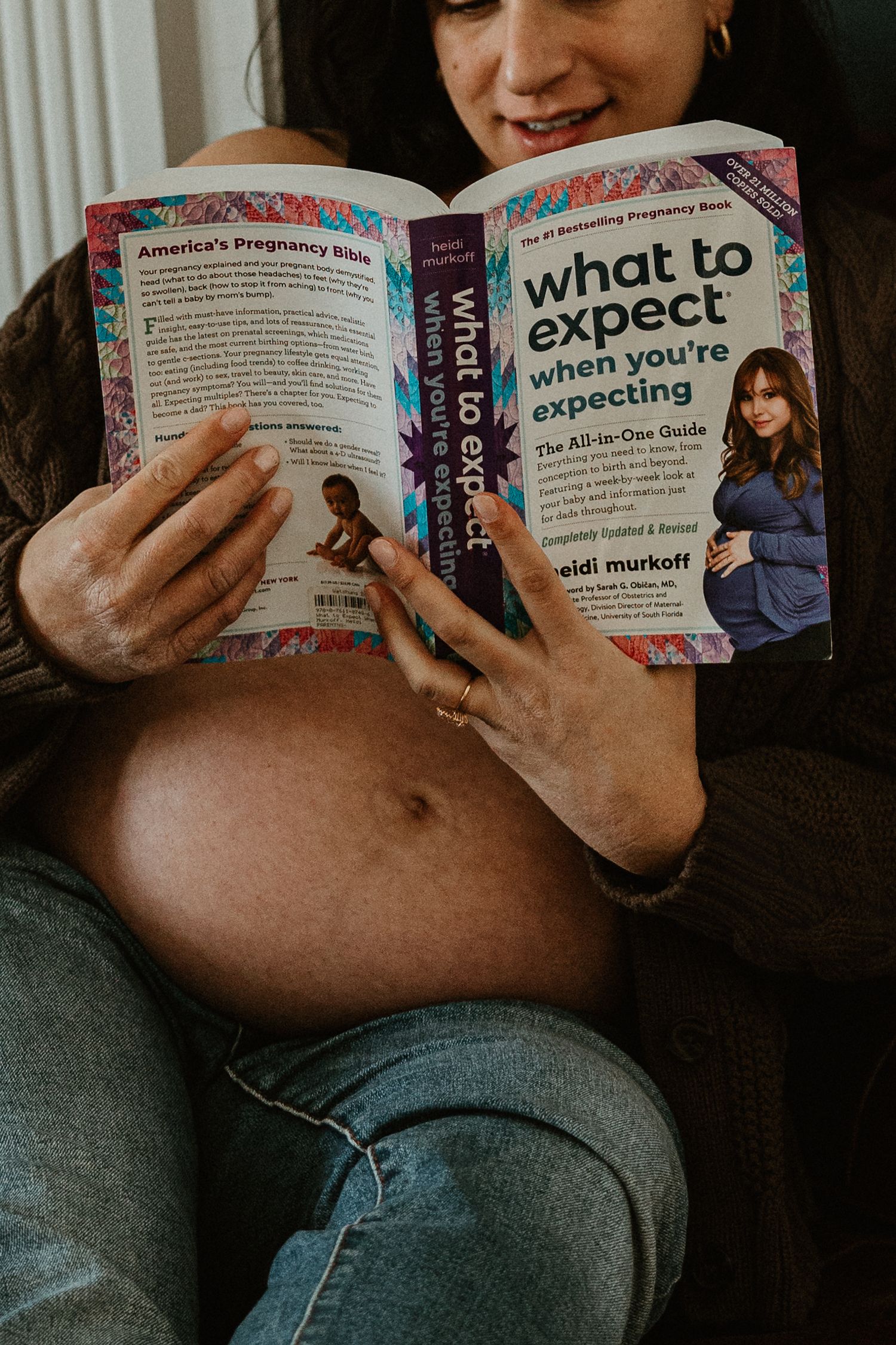Pregnant woman reads 'What to Expect When You're Expecting' book during maternity photoshoot.