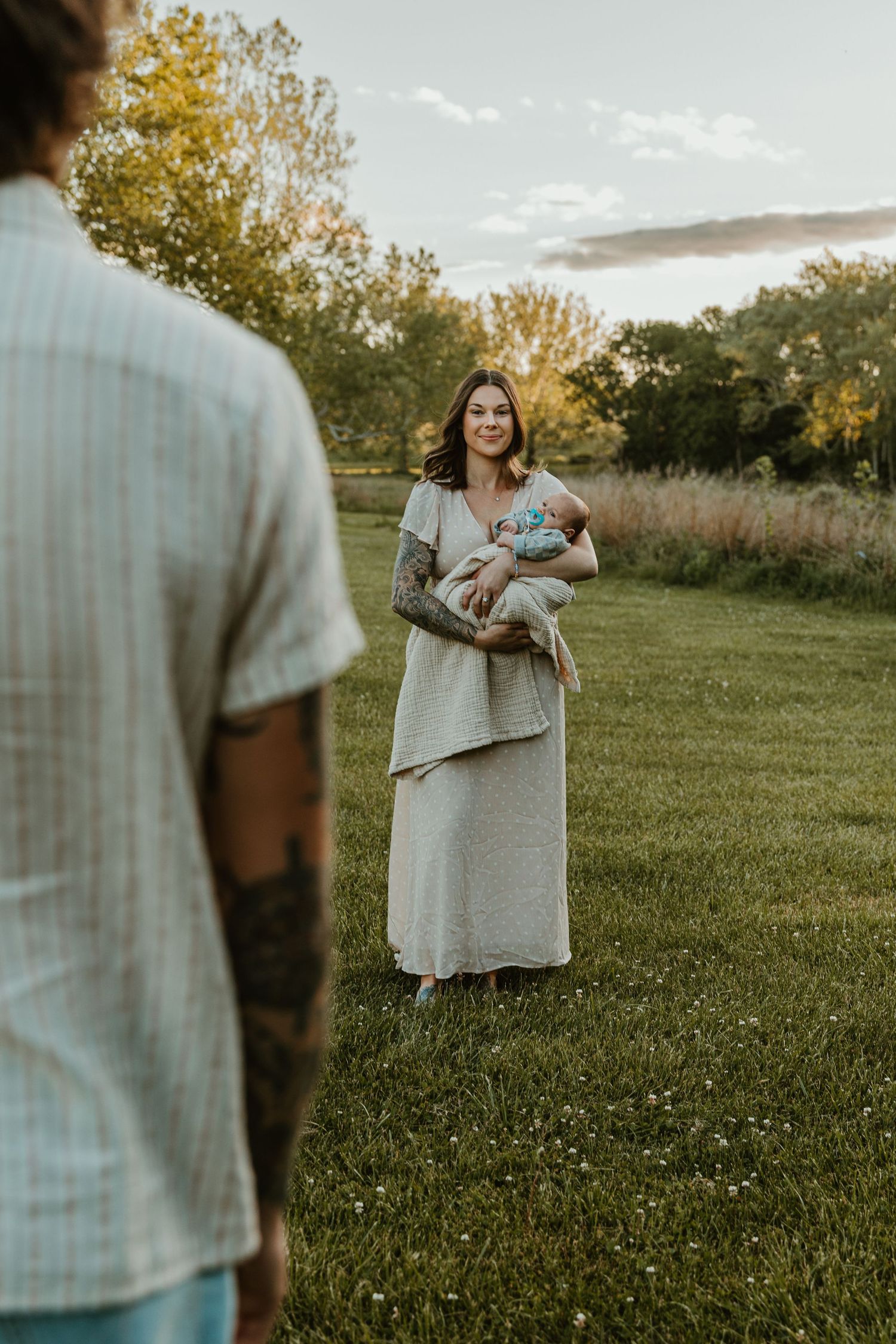 A tattooed mother holds her newborn baby in a field at sunset as her partner watches nearby.