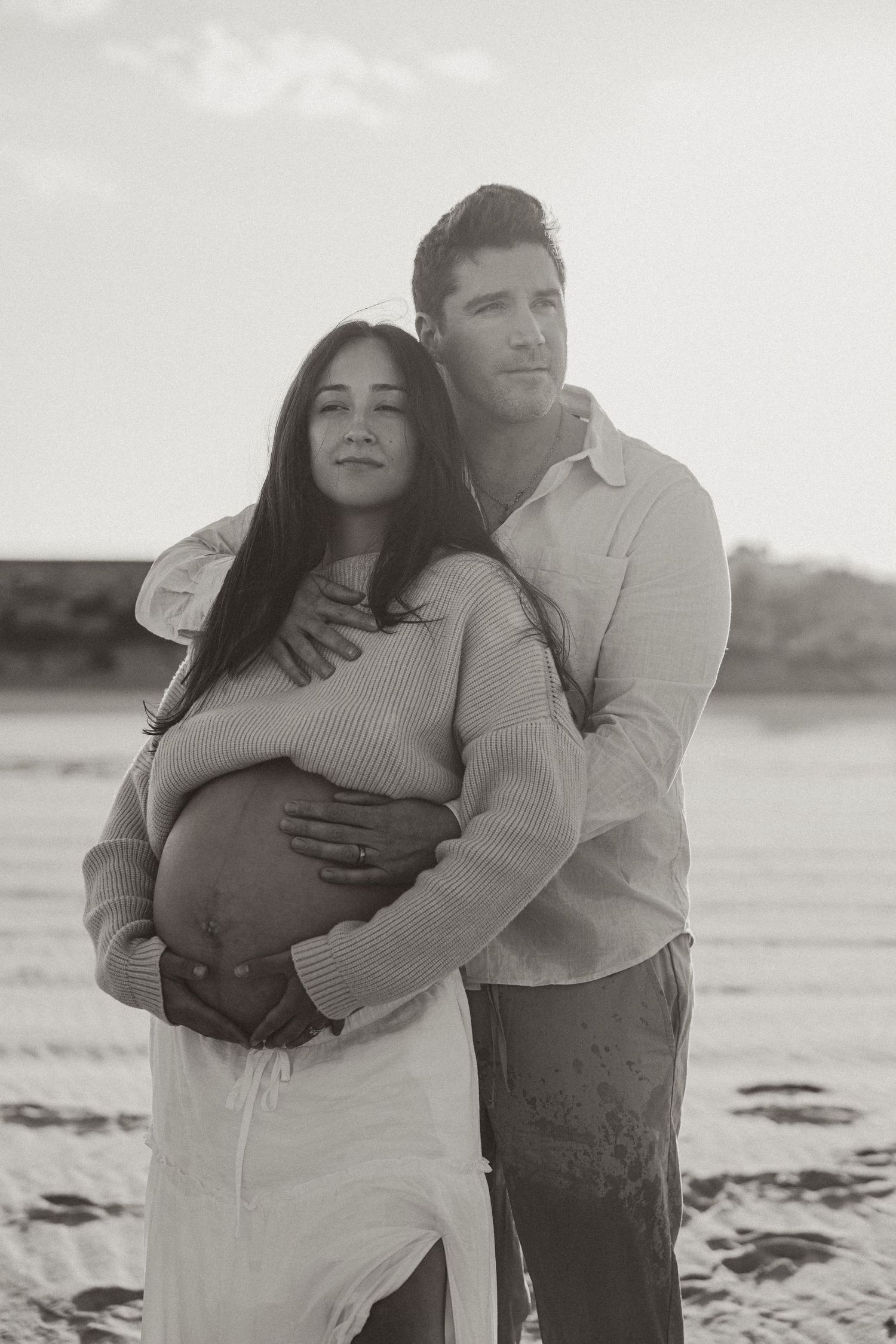 Black and white maternity photo of a couple embracing on a beach, man holding pregnant woman's belly.