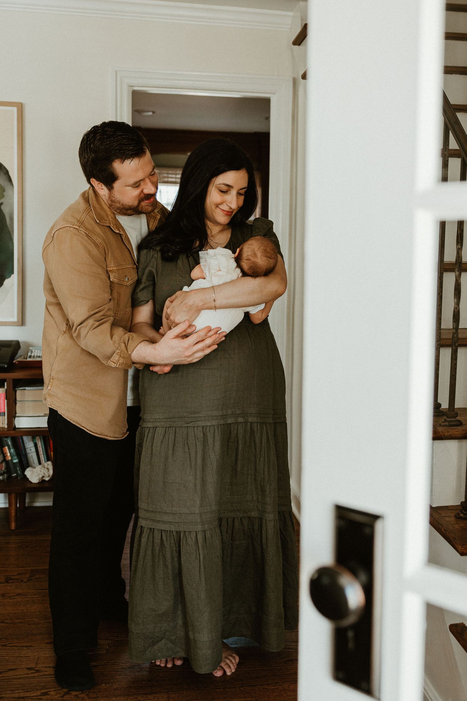 A couple smiles lovingly at their newborn baby, with the mother in an olive green dress holding the infant indoors.