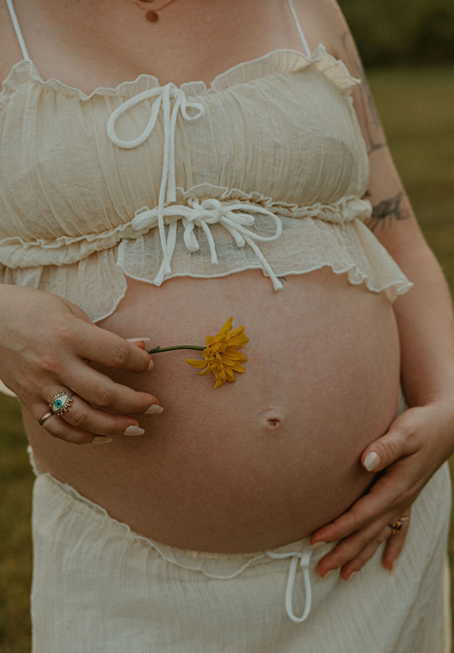 Pregnant woman in white crop top holding a yellow daisy against her bare baby bump outdoors.