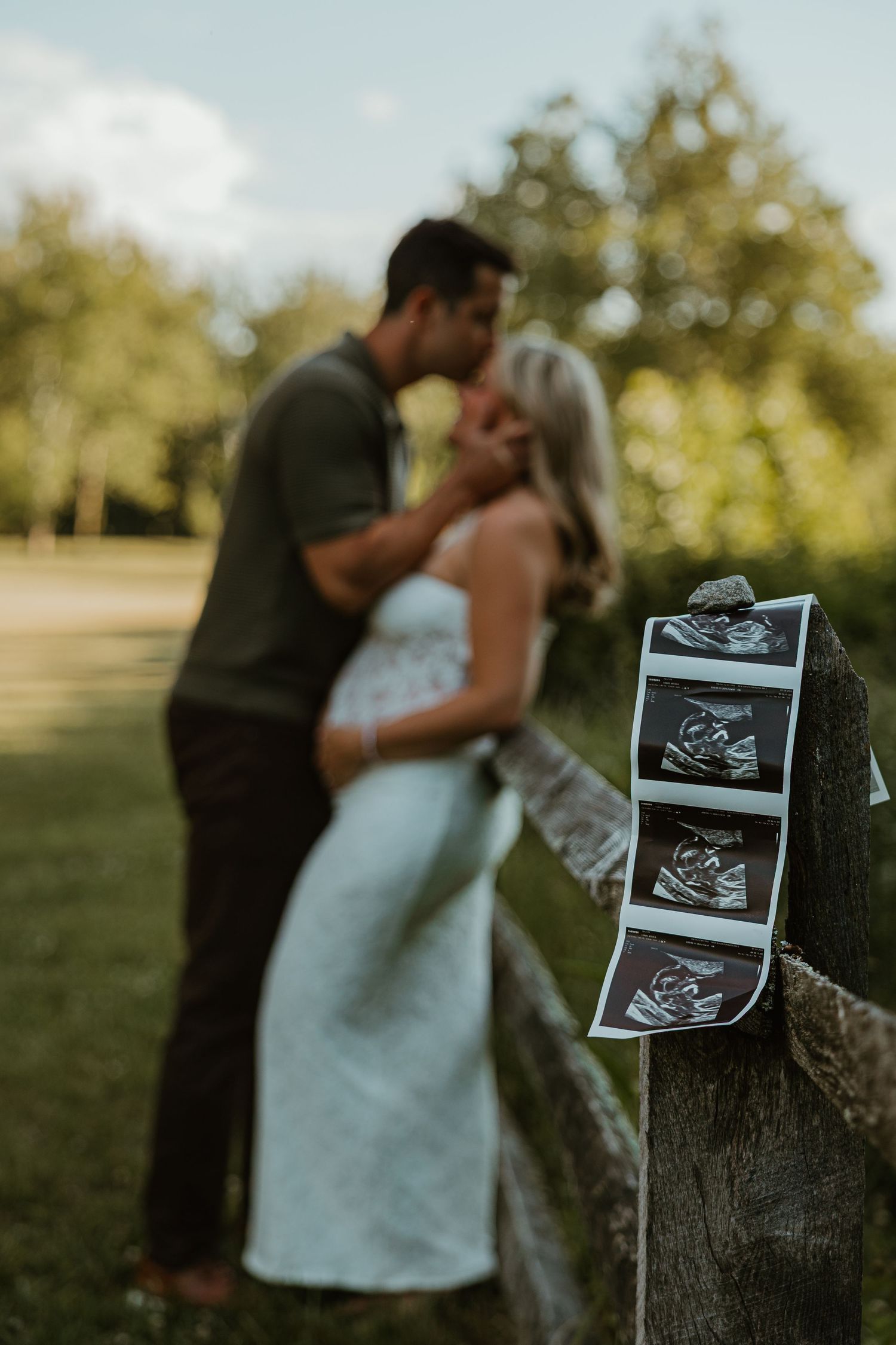 Couple kissing in a park during a pregnancy announcement photoshoot with ultrasound photos displayed on a wooden fence.