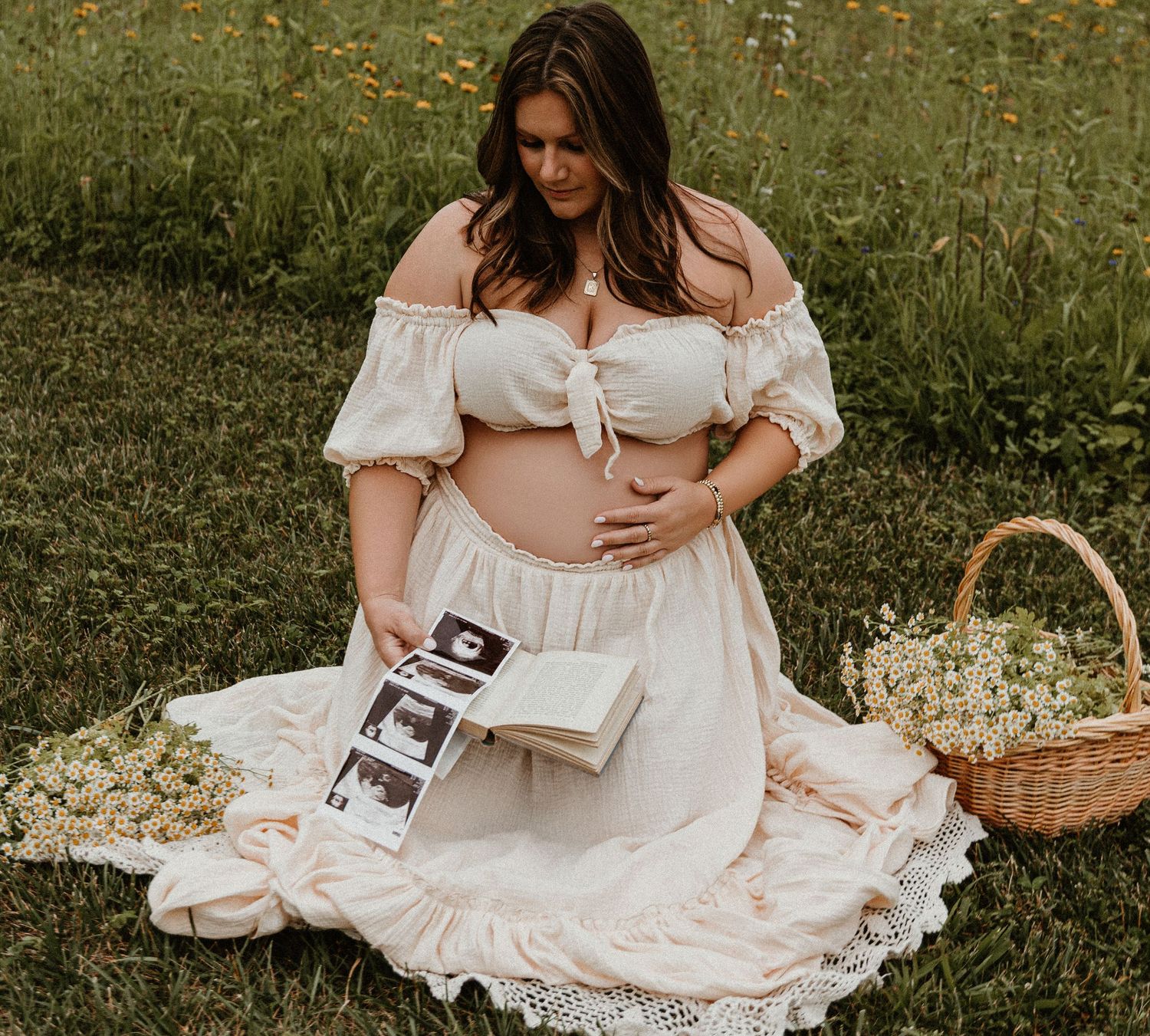 Woman in white dress sits in meadow with ultrasound photos and picnic basket.