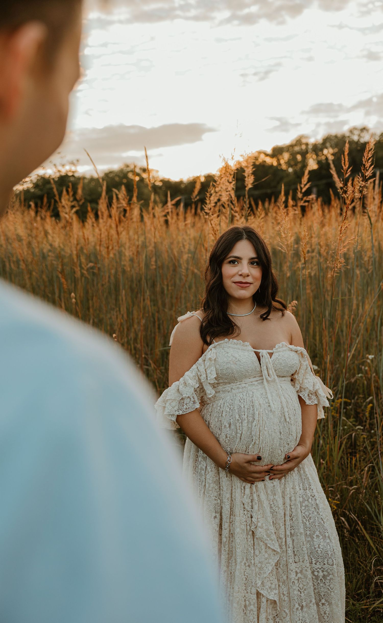Pregnant woman in white lace off-shoulder dress standing in golden field at sunset during maternity photoshoot.