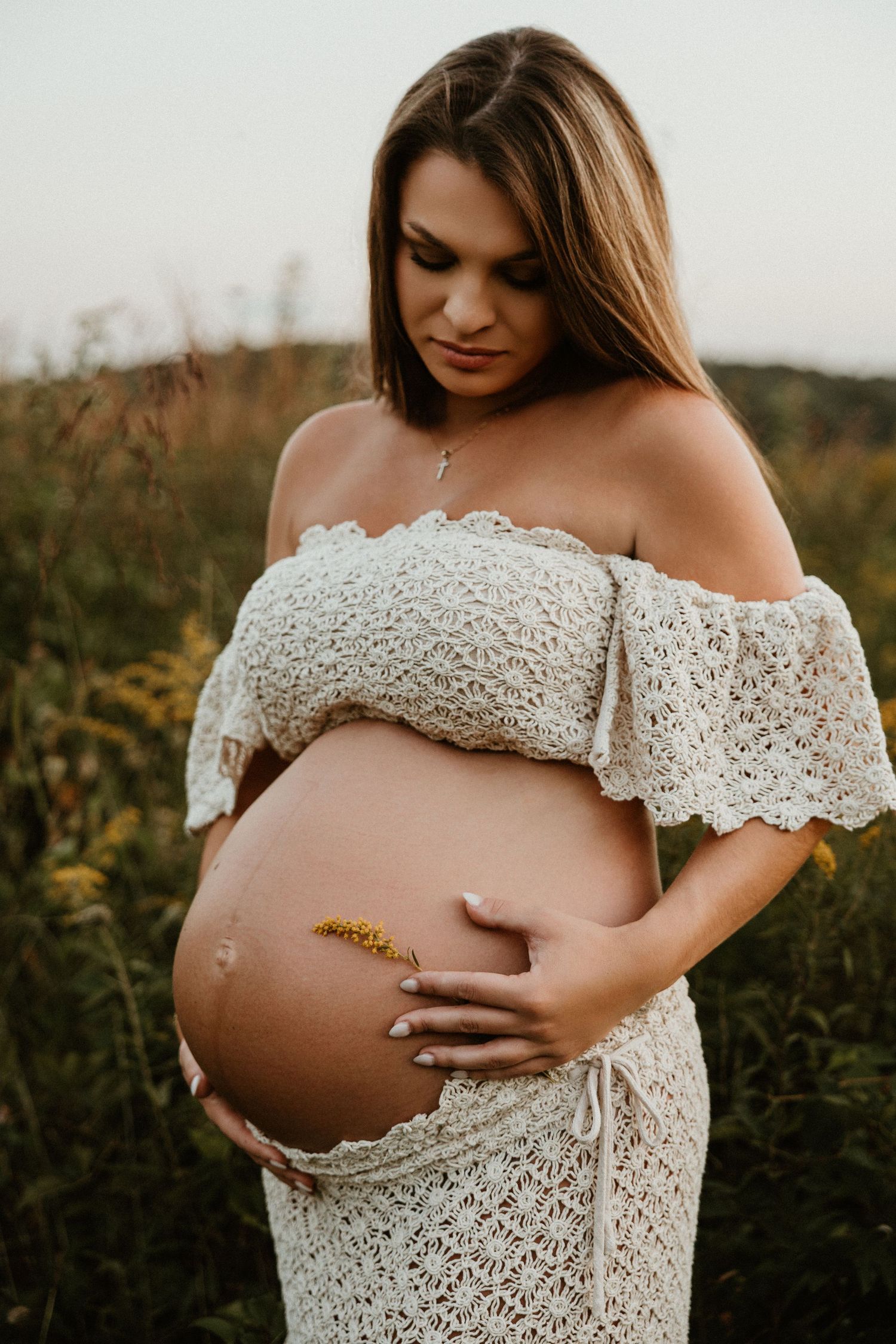 Close-up maternity portrait showing pregnant belly in off-shoulder white lace dress outdoors.