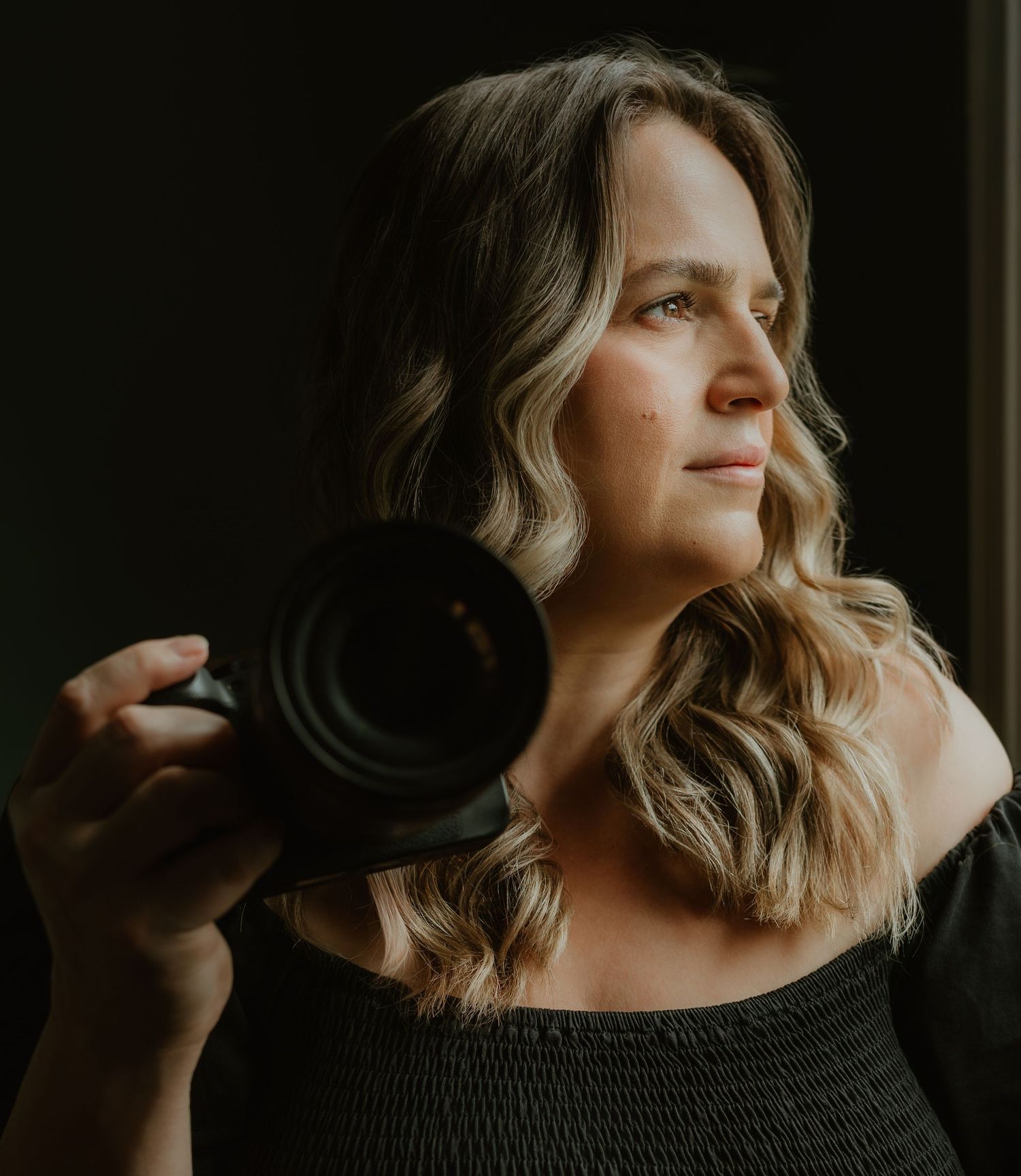A woman with blonde wavy hair holds a professional camera, gazing thoughtfully in moody studio lighting.