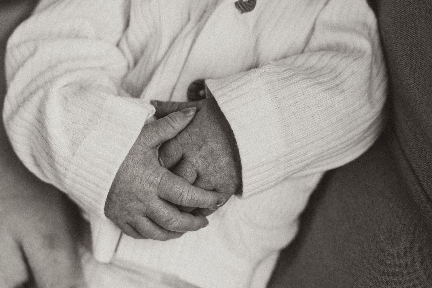Black and white photo of elderly person with clasped hands wearing a white knit sweater.