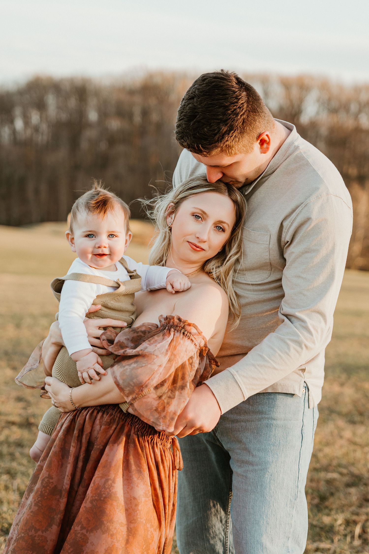 A young family poses outdoors at golden hour, mother holding baby while father embraces them both in a field.