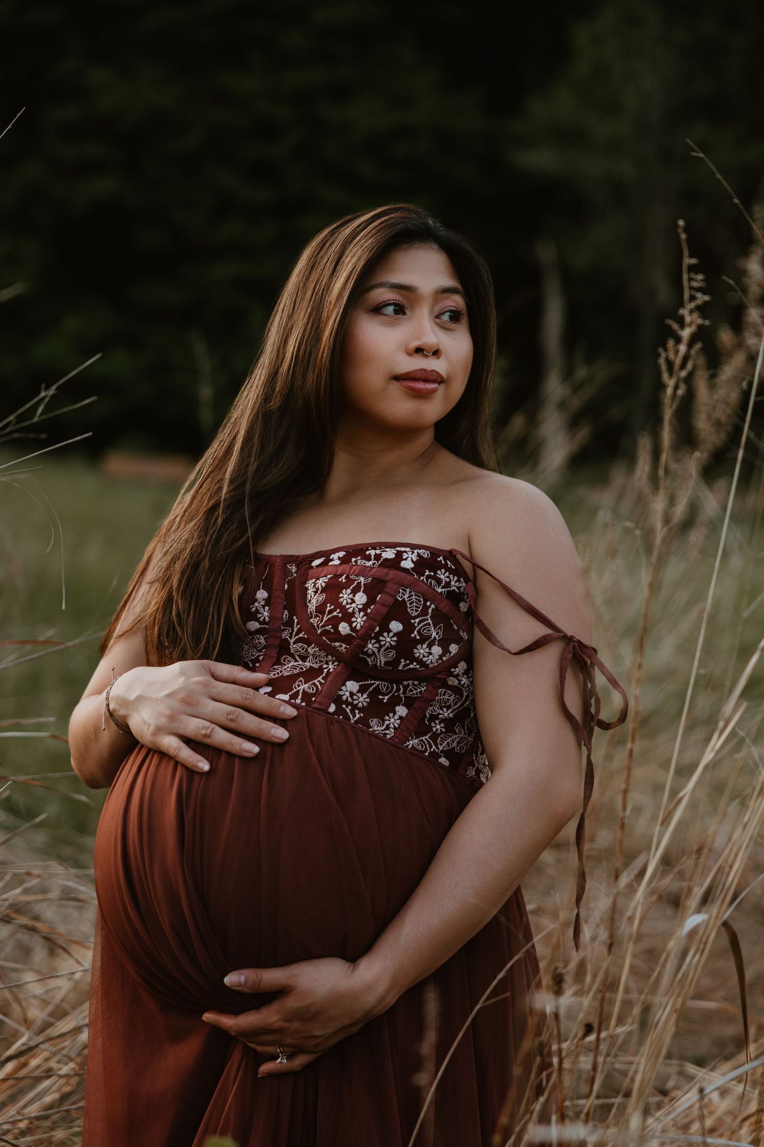 Pregnant woman in burgundy floral dress cradling baby bump outdoors in golden grass field with forest backdrop.