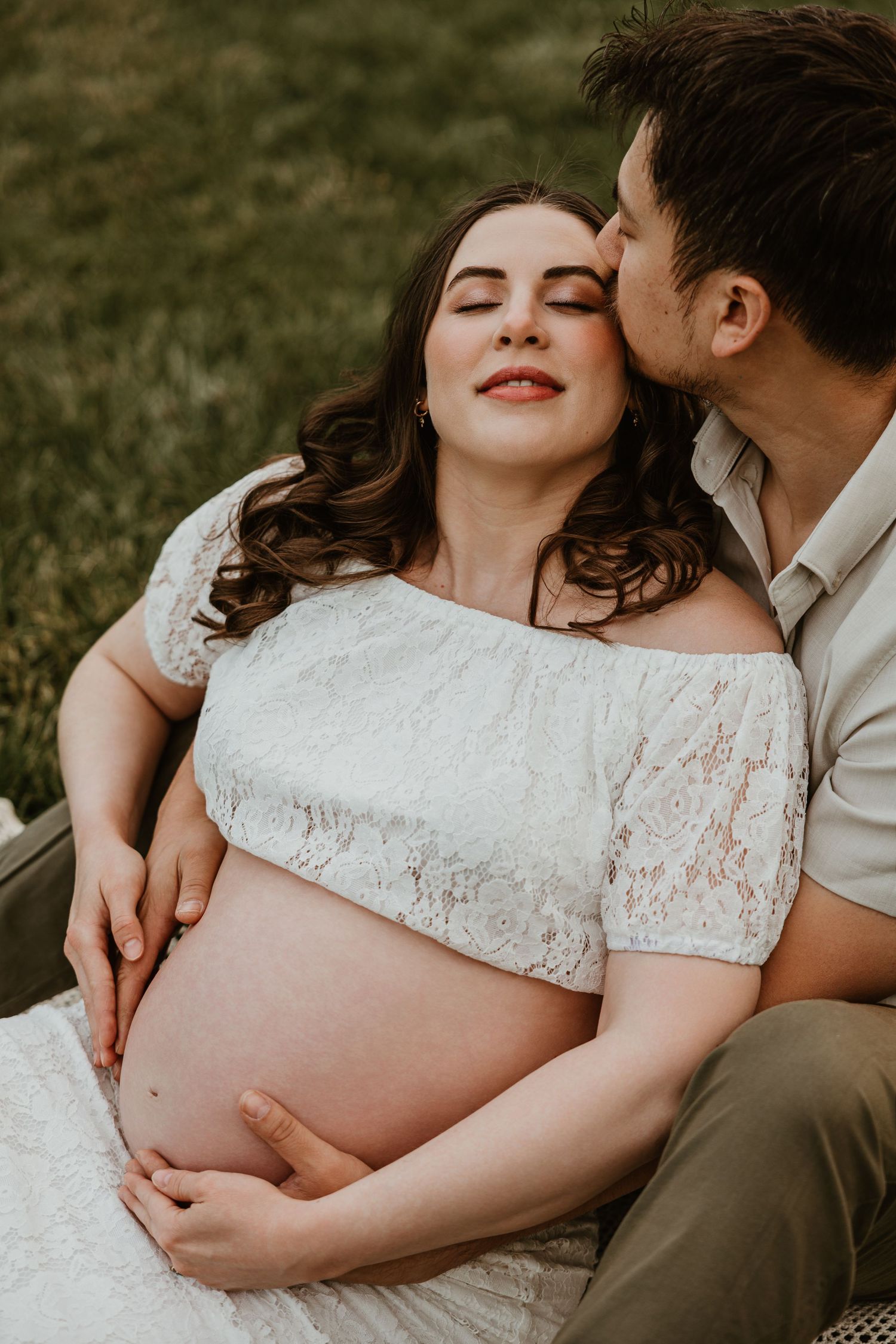 Pregnant woman in white lace dress sits outdoors as partner kisses her cheek, both cradling her baby bump.
