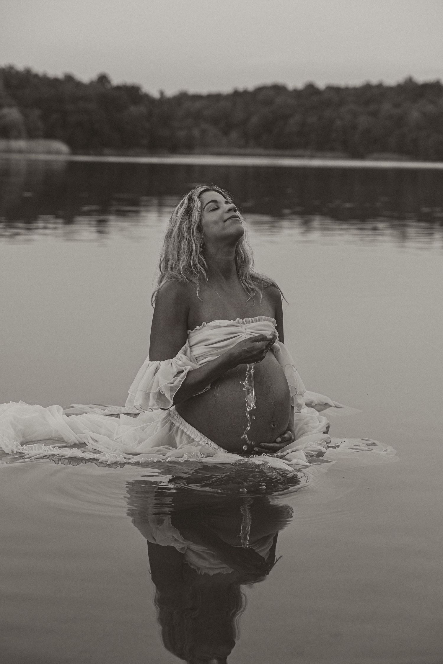 Black and white maternity photo of pregnant woman in white dress sitting in calm lake water.