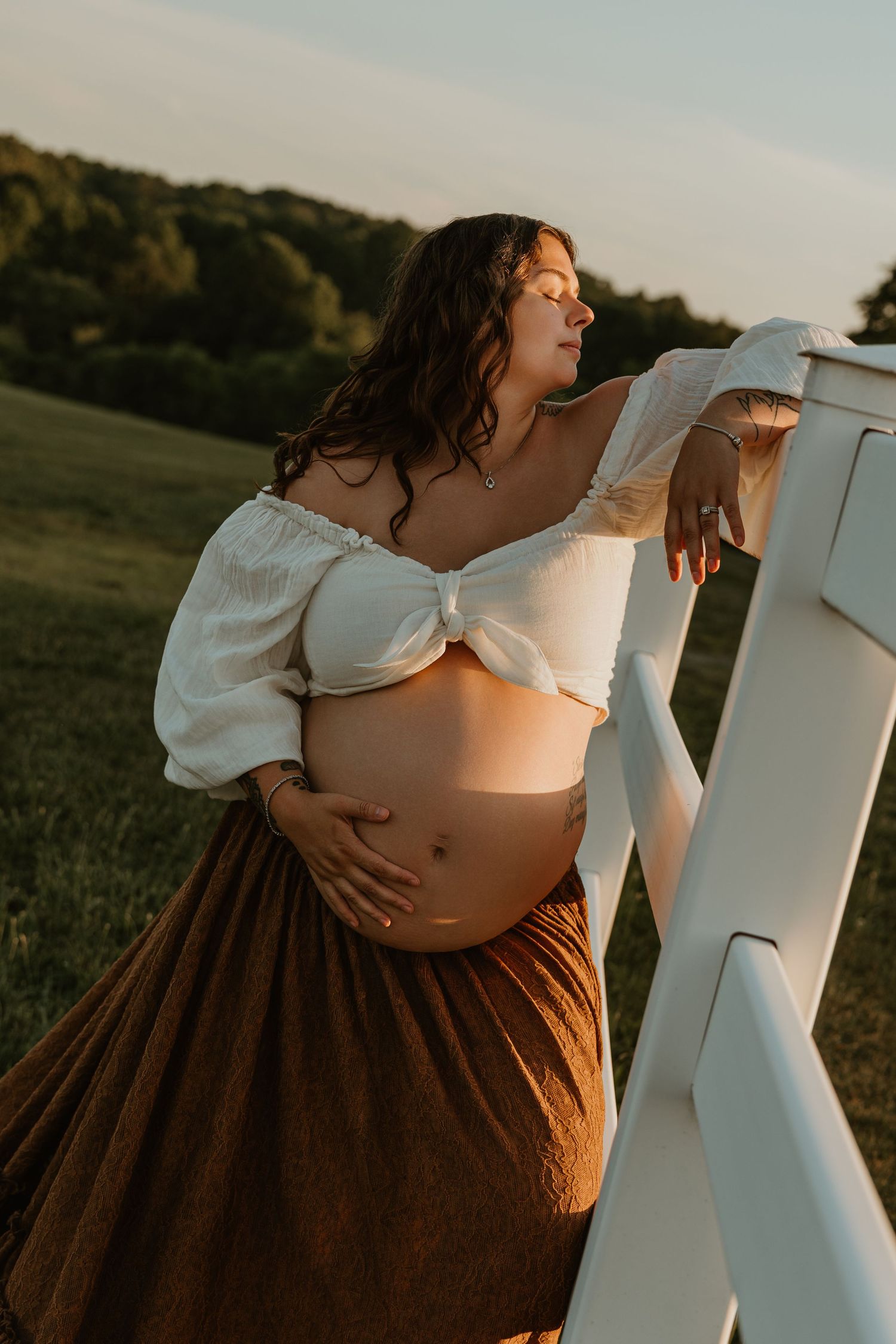 Pregnant woman in white crop top and brown skirt cradling baby bump outdoors at golden hour near white fence.
