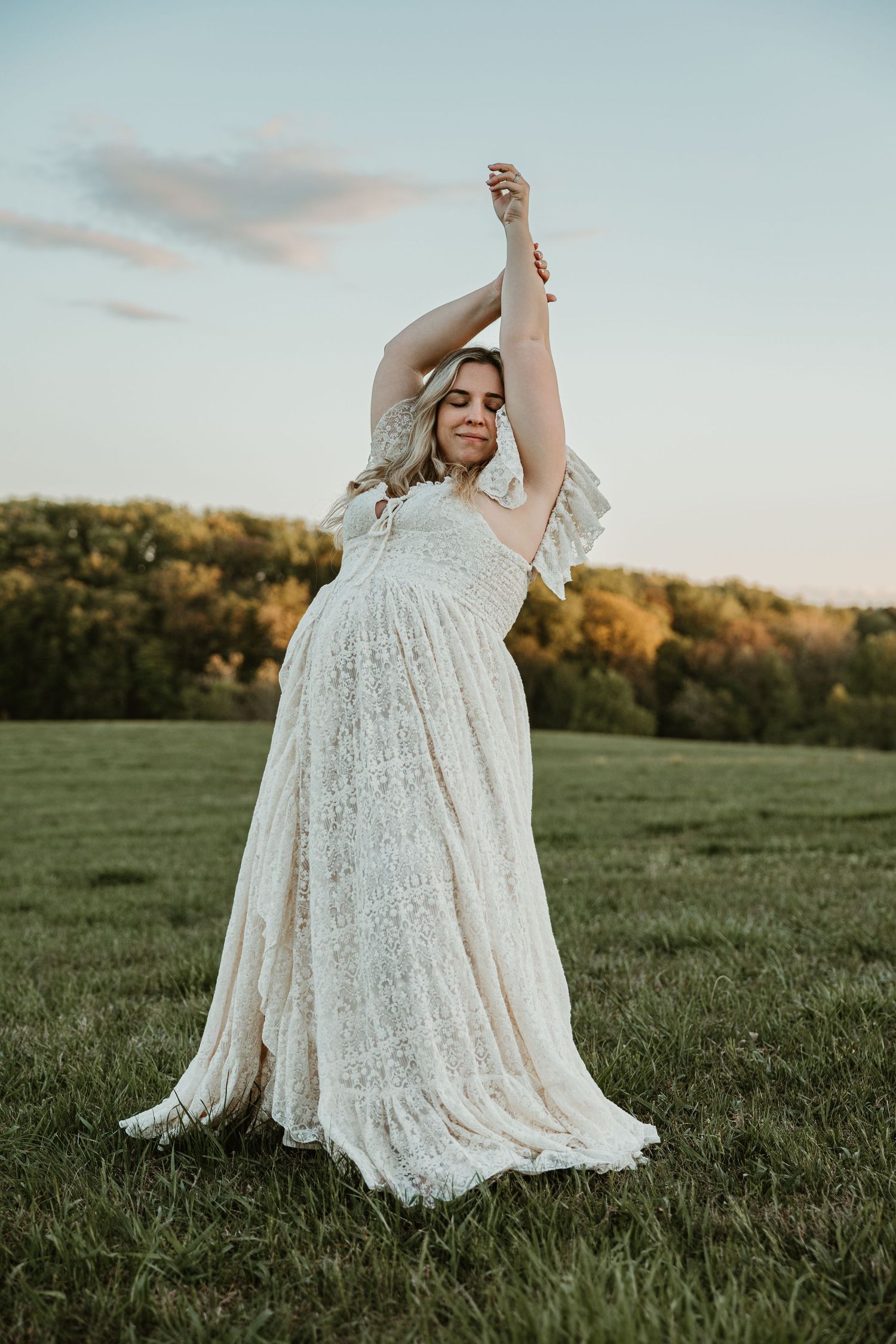 Pregnant woman in white lace boho gown dancing in green field at golden hour sunset.