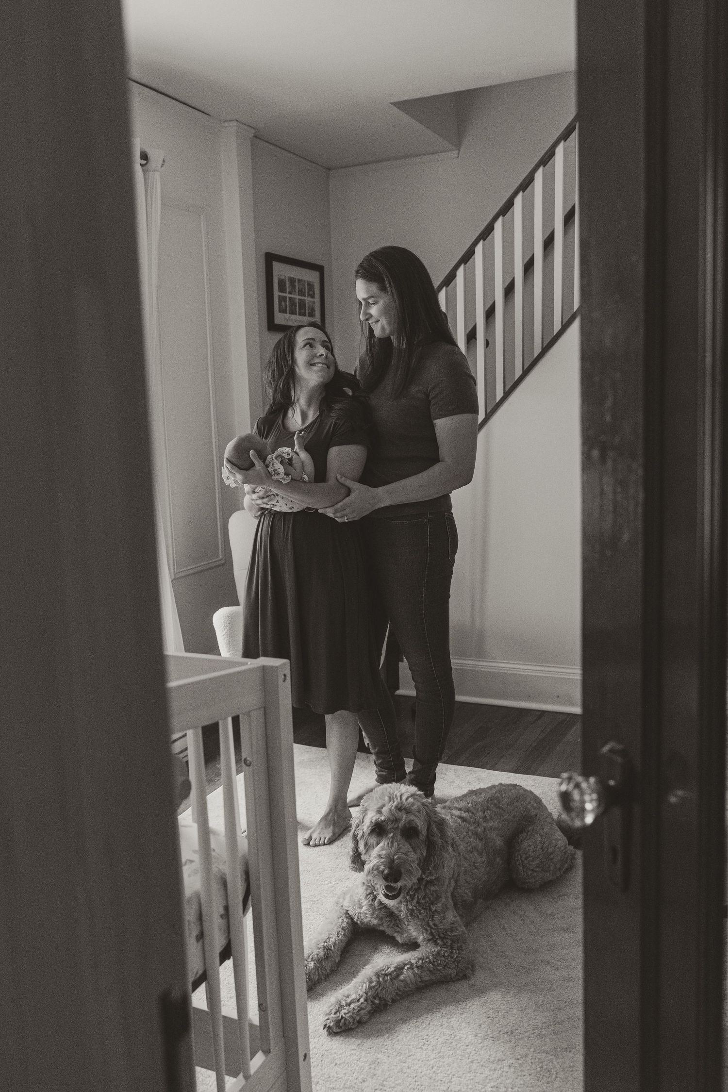Two women holding a newborn baby in a home hallway with a fluffy dog resting on the floor nearby.