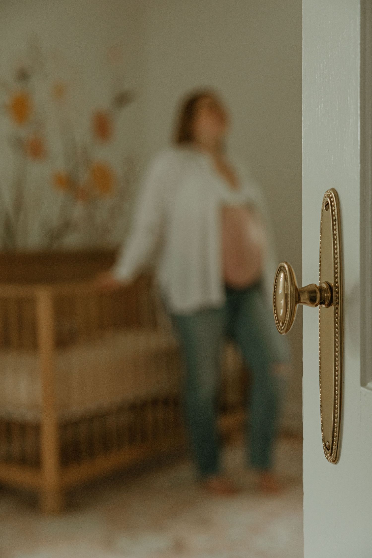 Pregnant woman standing in a nursery room viewed through a doorway with a vintage gold door knob in focus.