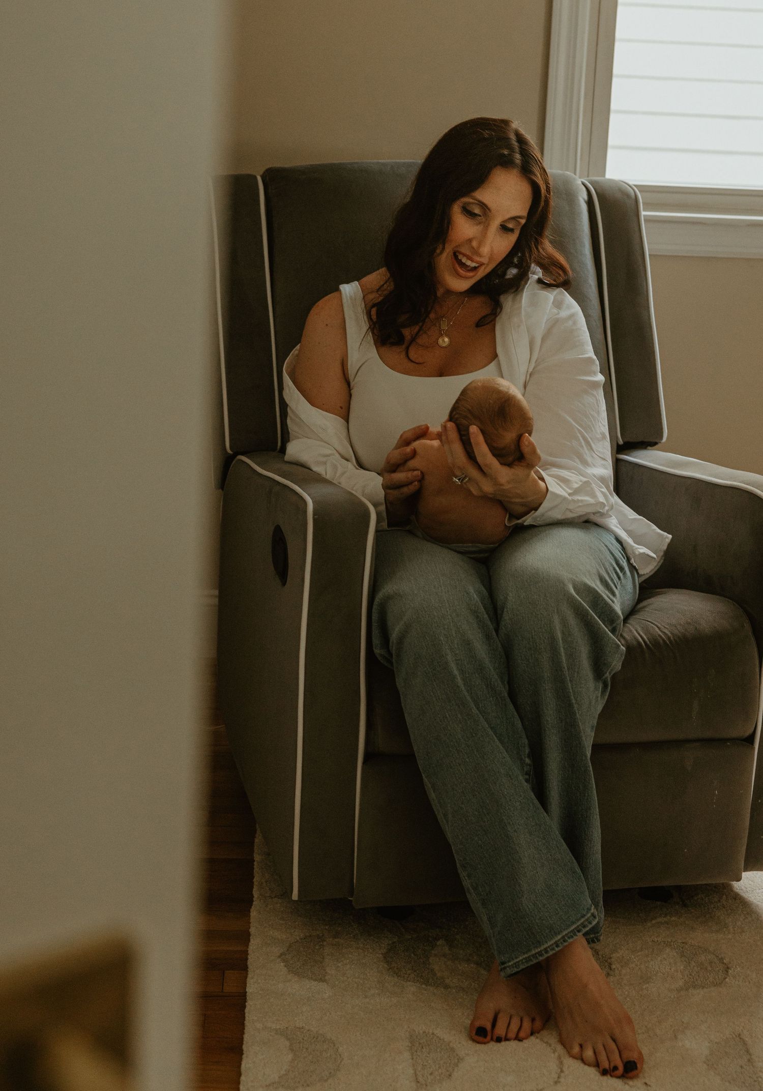 A mother sits in a gray recliner chair, smiling down at her newborn baby in a warmly lit nursery room.