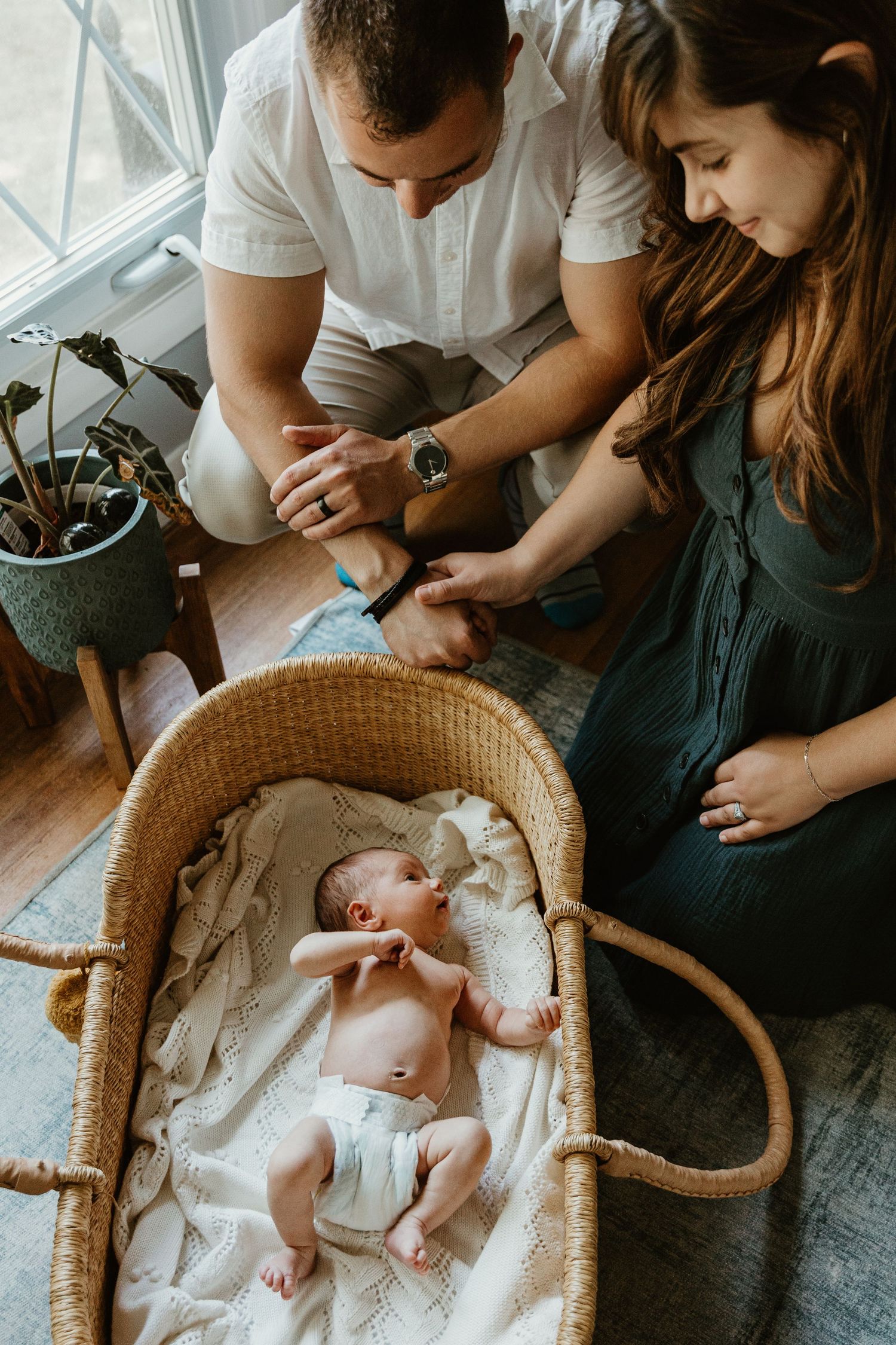 Newborn baby resting in woven bassinet while parents watch from above.