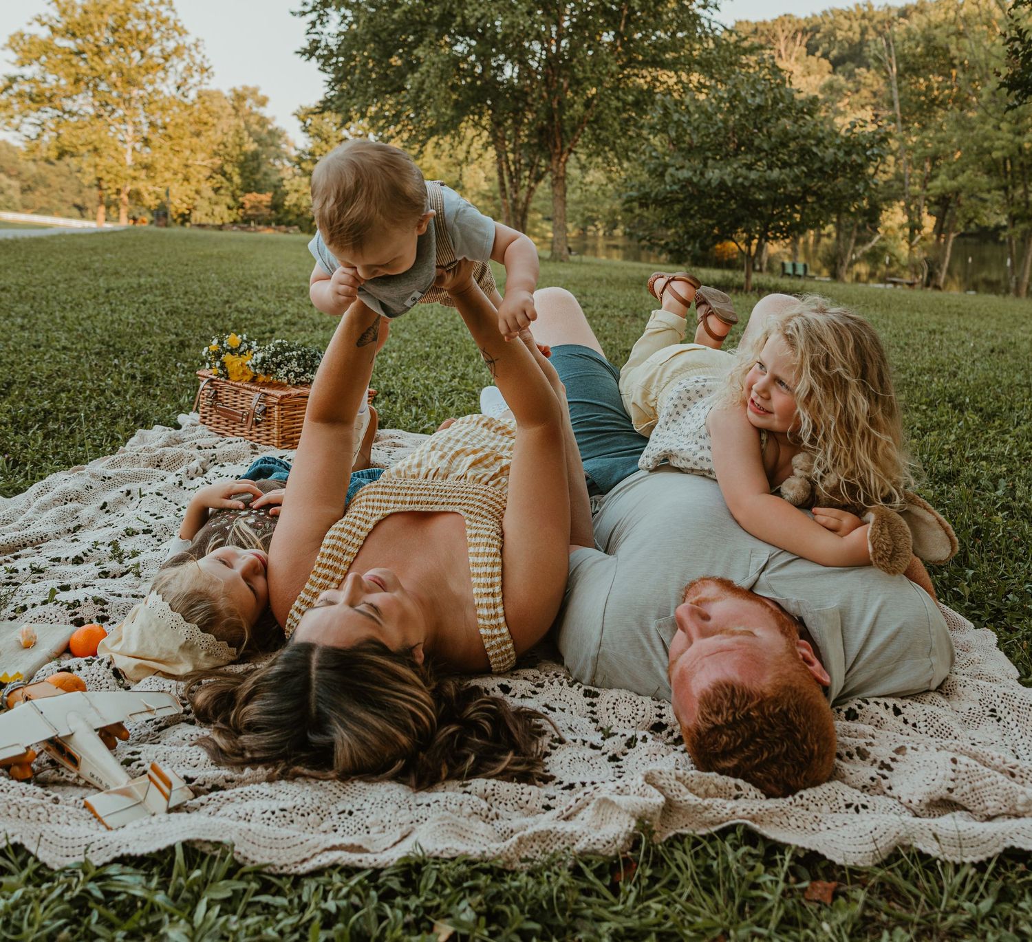 Family enjoying a sunny picnic on a blanket in the park, parents lying down while playing with two young children outdoors.