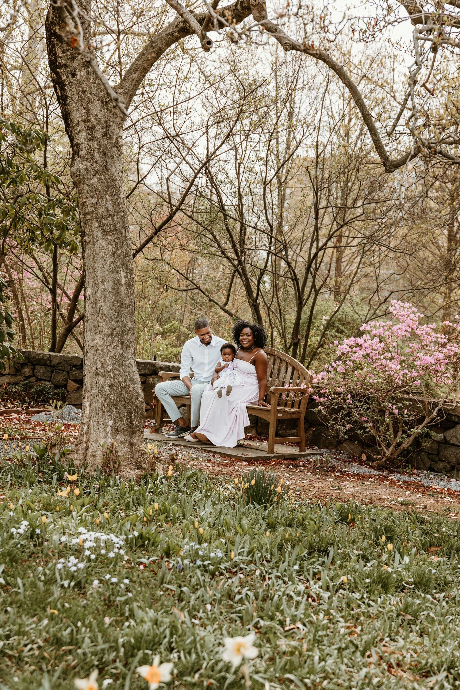 Family sitting on wooden bench under blooming spring trees in garden setting.