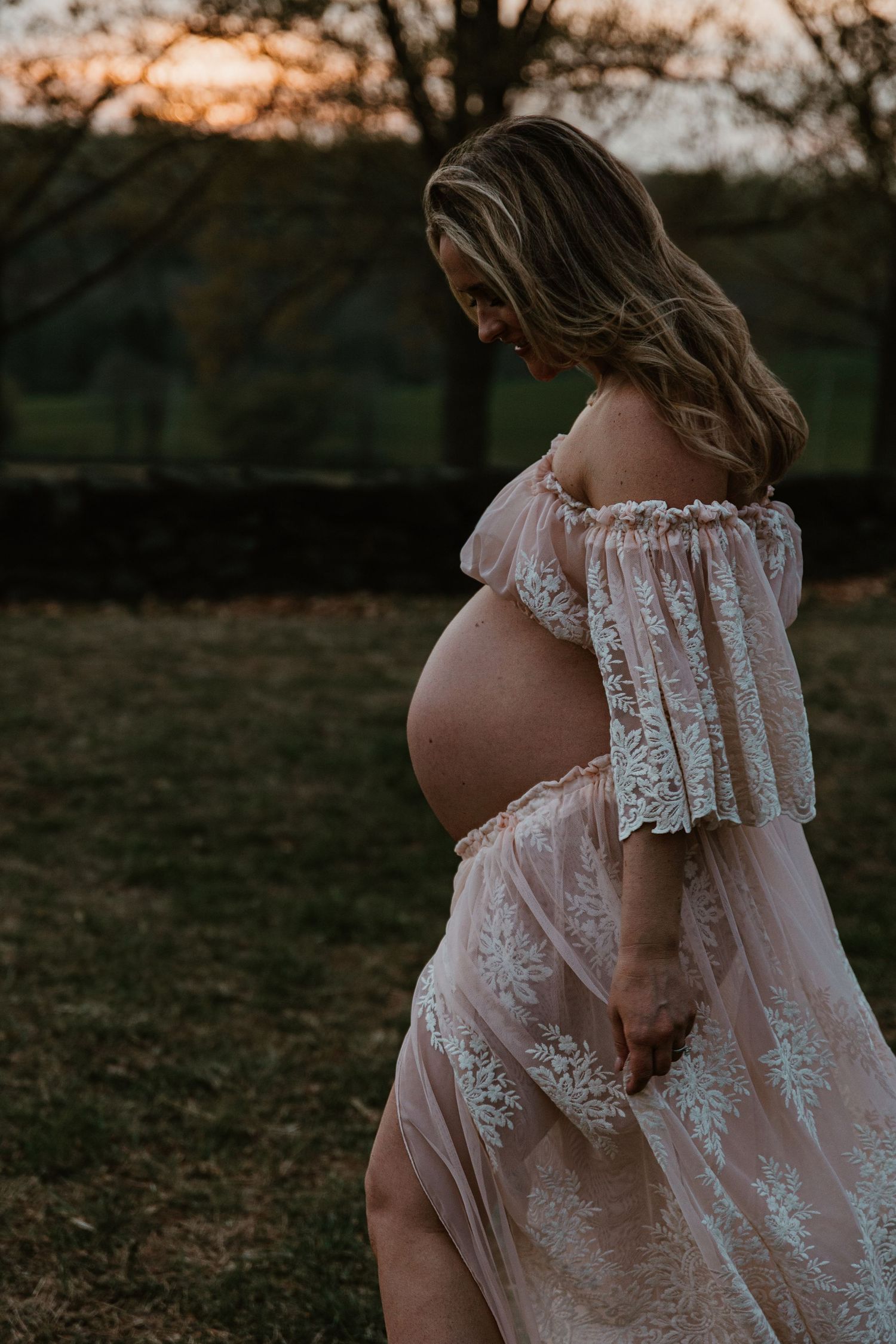 Side view of pregnant woman in flowing lace dress standing in garden at sunset.