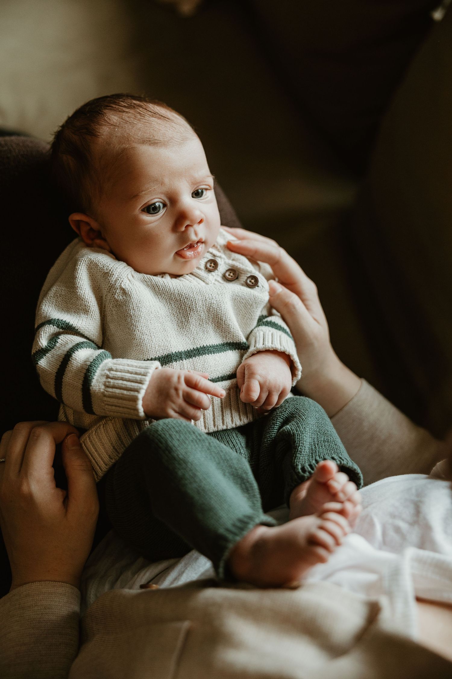 Infant in cream colored sweater and green pants lying on white bedding in moody lighting.